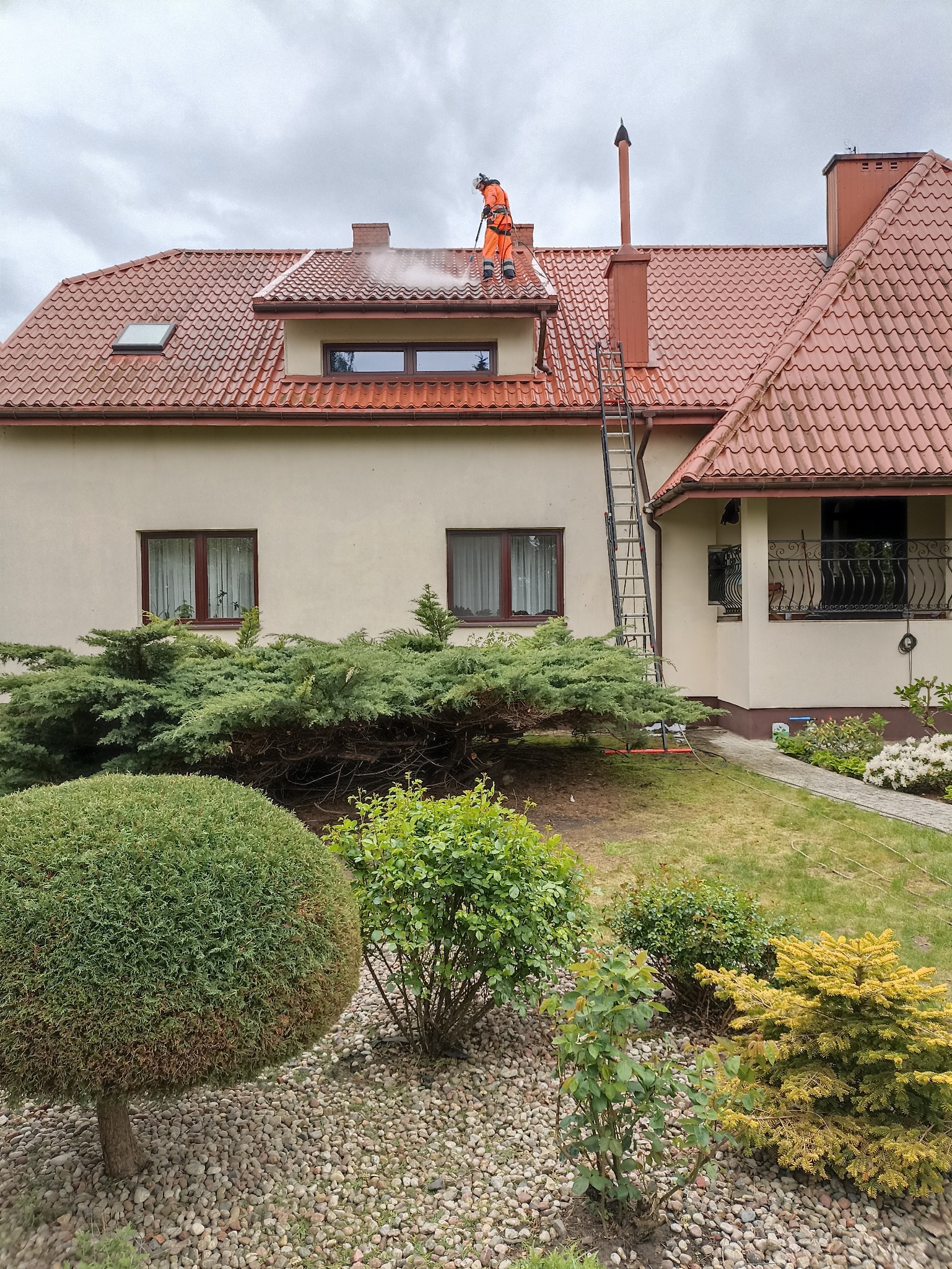 Pracownik w kombinezonie myje dach z czerwonej dachówki domu jednorodzinnego za pomocą myjki ciśnieniowej. Widoczna drabina oparta o budynek i zadbany ogród.