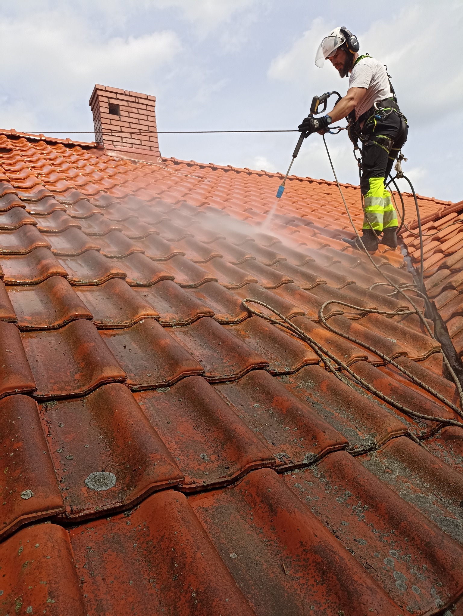 Mycie ciśnieniowe dachówki w Płocku. Pracownik w uprzęży czyści dach z mchu, używając lancy. Widoczny komin i fragment dachu przed i po czyszczeniu.