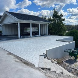 brukar - Jasnoszara kostka brukowa na podjeździe w Lublinie, prowadząca do garażu z przyczepą. Widok na jezioro i błękitne niebo z chmurami. W tle dom z ciemnym dachem.