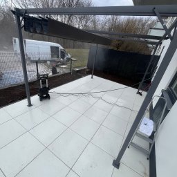 brukar - Nowoczesny taras z jasnych płyt, zadaszony pergolą w kolorze grafitowym. Widoczna myjka ciśnieniowa, drabina i częściowo zwinięte zadaszenie. W tle ogrodzenie i biały bus.