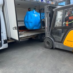 brukar - Wózek widłowy Jungheinrich ładuje niebieski zbiornik do białego busa. W tle widoczne materiały budowlane na regałach.