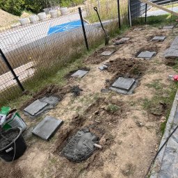 brukar - Ustawianie betonowych płyt fundamentowych w ziemi z zaprawą, z widocznym sznurkiem do poziomowania, wiadrem cementu i łopatą przy siatce ogrodzeniowej, w tle parking z niebieskim oznaczeniem.