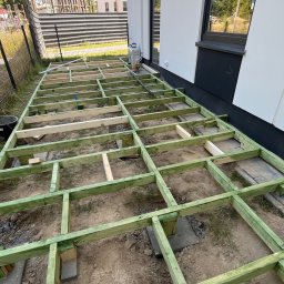 brukar - Zielona, impregnowana drewniana konstrukcja nośna pod taras, ułożona na gruncie obok nowoczesnego budynku z białej elewacji i ciemnymi oknami. Widoczne wsporniki z bloczków betonowych.