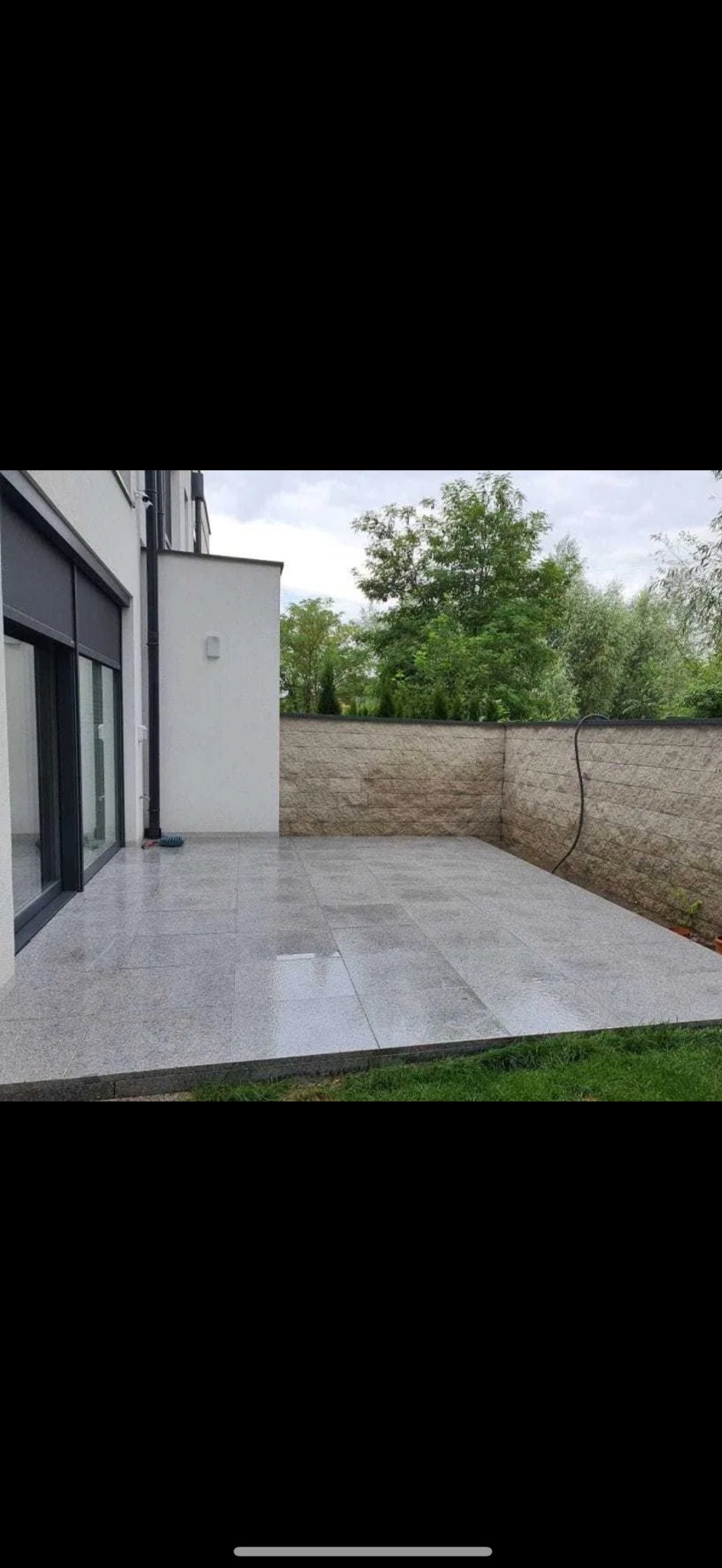 Wyłożony jasnoszarymi płytami taras przydomowy, z widoczną ścianą z kamienia i fragmentem budynku z dużym oknem. Na tarasie leży niebieska szczotka.