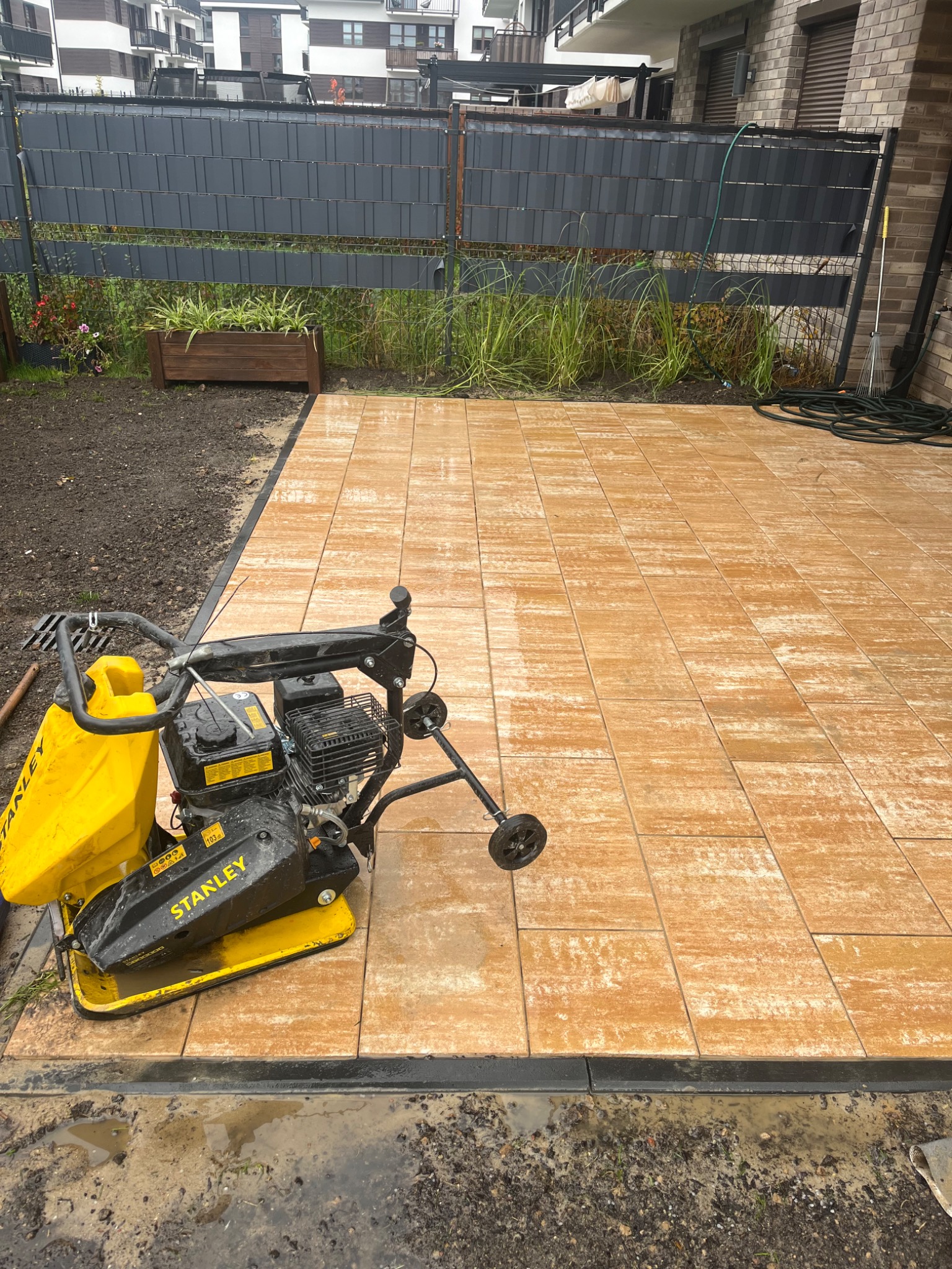 Ułożona kostka brukowa w odcieniach beżu, z widoczną ubijarką Stanley. Tło: nowoczesny budynek mieszkalny i ogrodzenie z roślinnością.