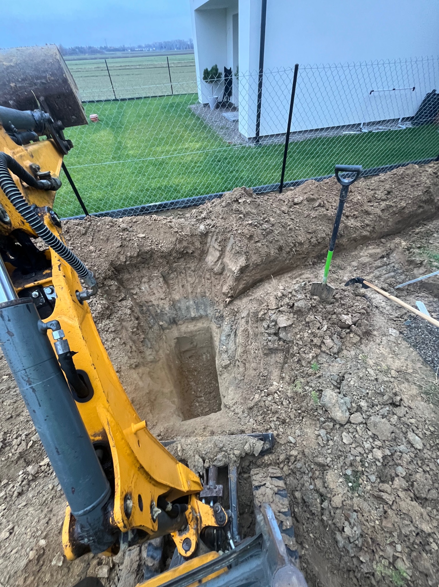 Żółta koparka przy wykopie w ziemi, obok ogrodzenie z siatki i budynek. Widoczne narzędzia: łopata i kilof. Prace ziemne w toku, teren przygotowywany pod instalacje.