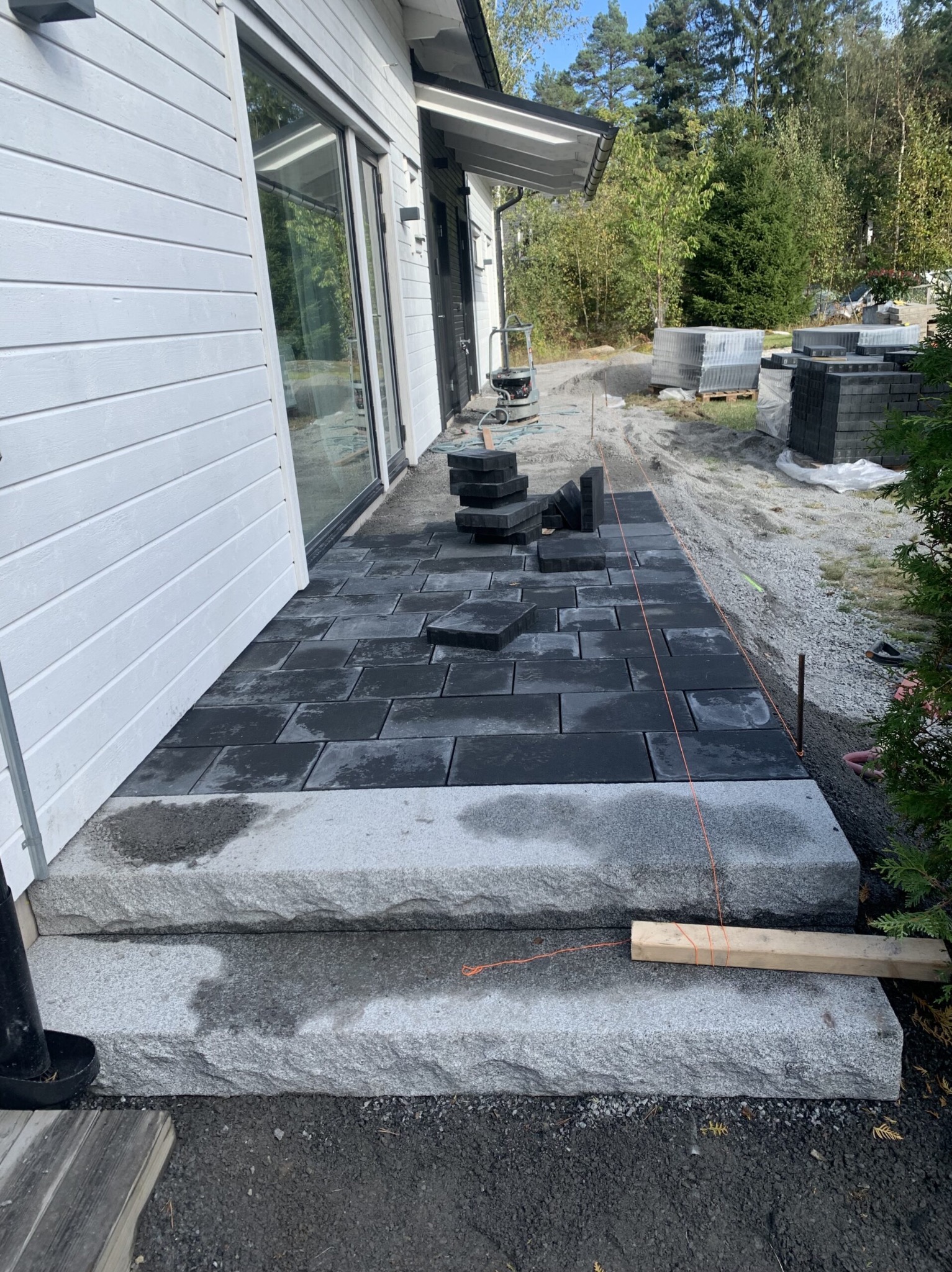 Układanie ciemnej kostki brukowej na posesji przy domu z białymi panelami. Widoczne granitowe schody wejściowe i palety z kostką w tle.