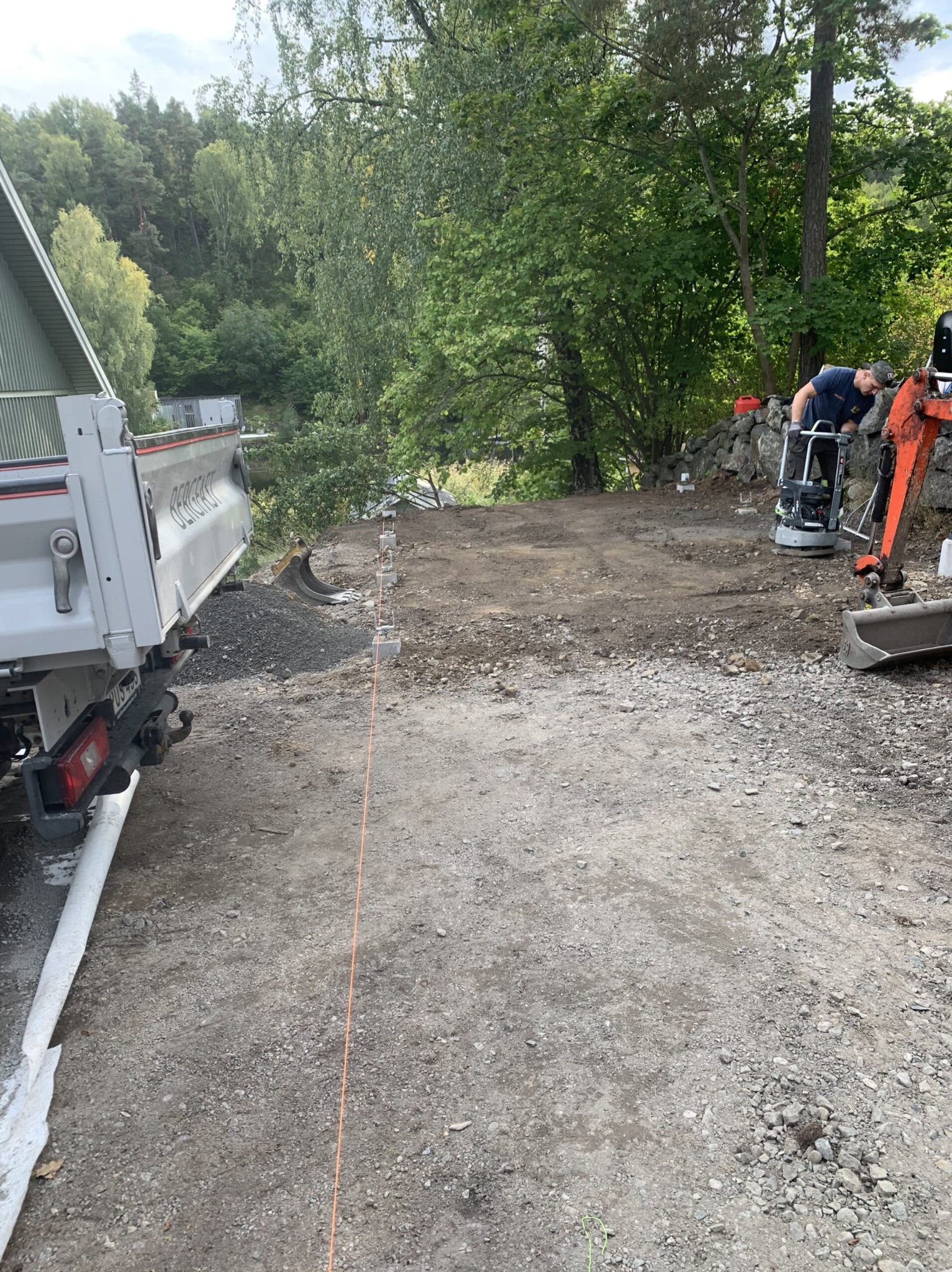 Prace ziemne: zagęszczarka w akcji, wyrównywanie terenu pod budowę. W tle widoczny wywrotka z materiałem i wytyczone granice przyszłej inwestycji.