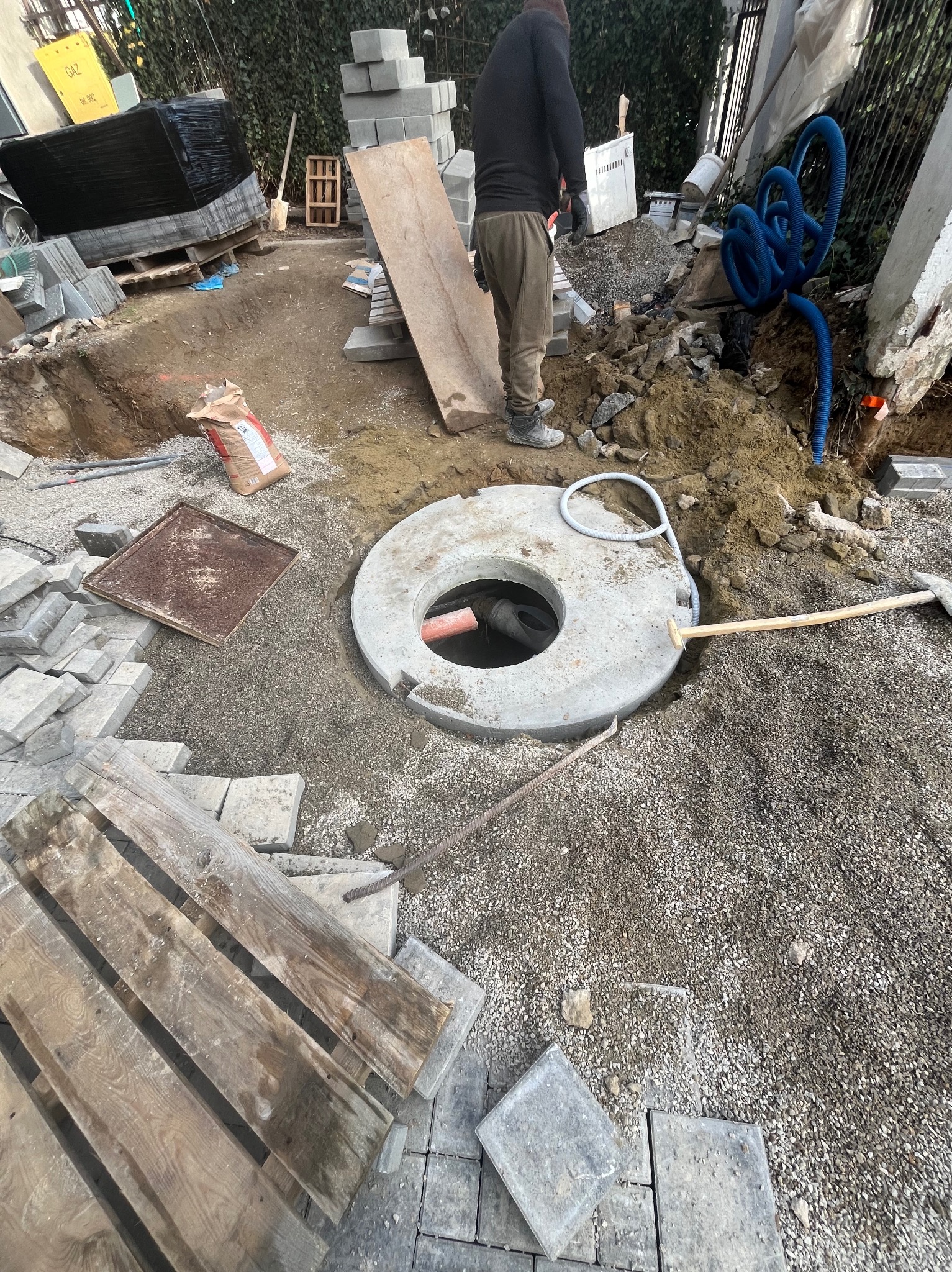 Przygotowanie terenu pod kostkę brukową: wykop z betonowym kręgiem, podsypka żwirowa, paleta z kostką, widoczny pracownik i narzędzia. Prace ziemne w toku.