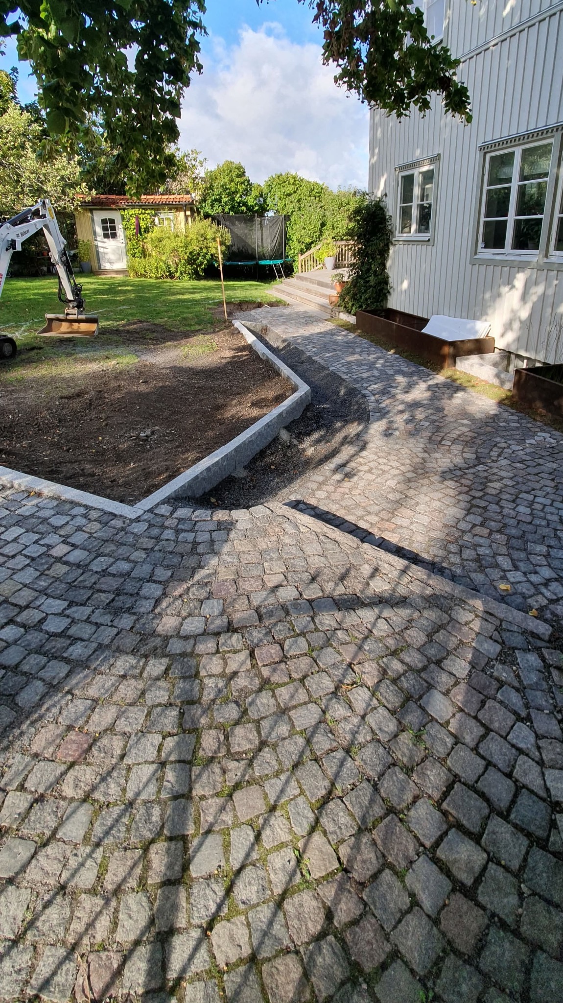 Granitowa ścieżka w ogrodzie z mini koparką w tle. Częściowo ułożona kostka brukowa z obrzeżami. Widok na dom i zieleń. Słoneczny dzień.