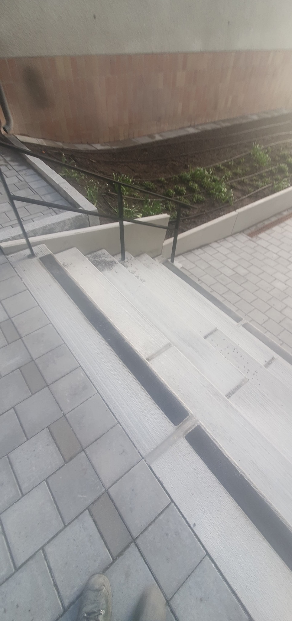 Schody z kostki brukowej z odpływem liniowym, zakończone rabatą. Widoczne fragmenty buta. Ujęcie z góry, podkreślające detale wykończenia i precyzję ułożenia.