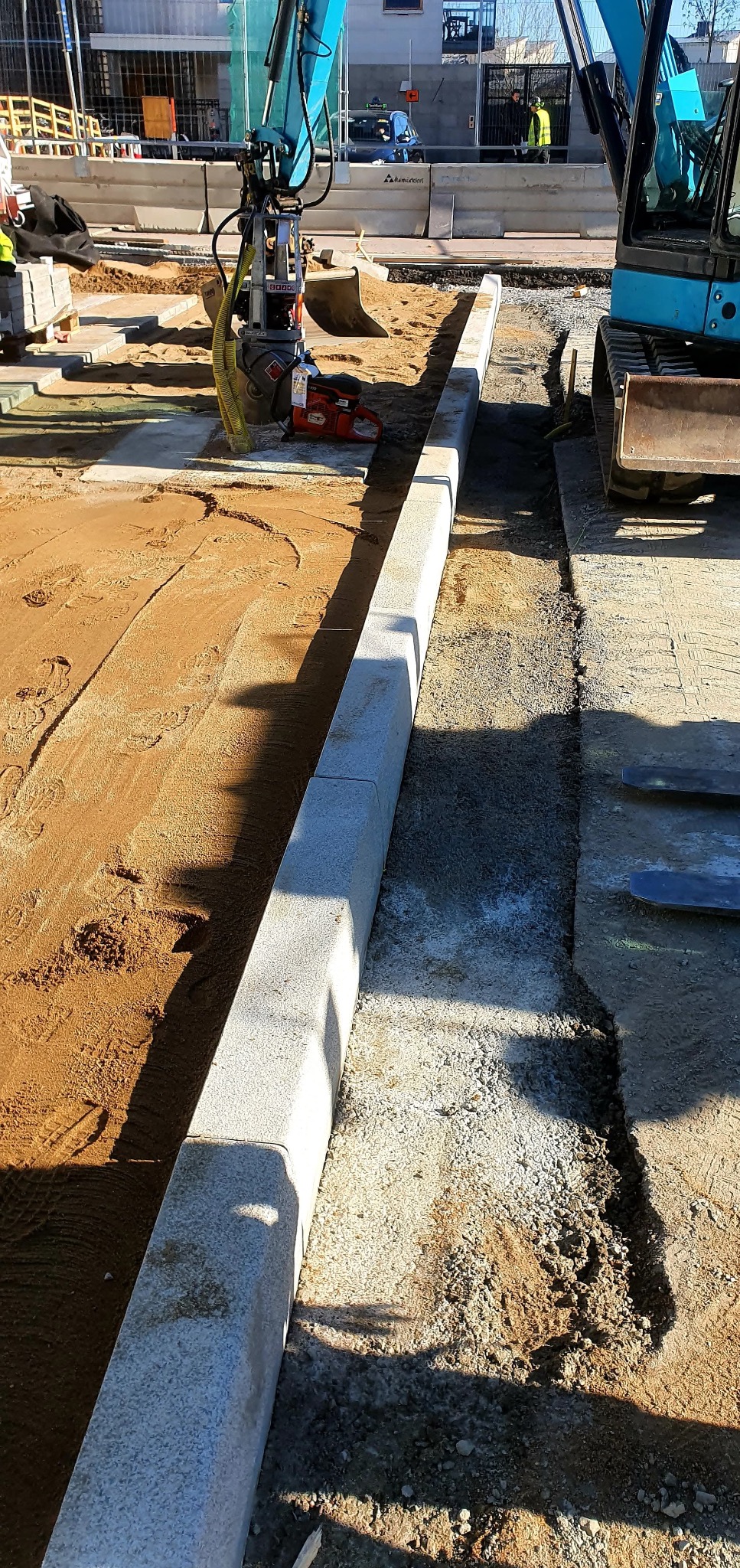 Montaż obrzeży chodnikowych z użyciem mini koparki i zagęszczarki. Obrzeża z jasnego kamienia oddzielają piasek od podłoża z kruszywa.