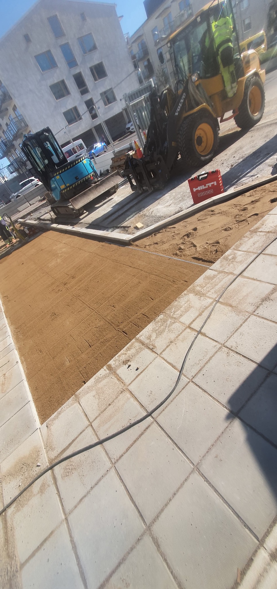 Przygotowany teren pod kostkę brukową z użyciem mini koparki i walca. Widoczny fragment ułożonej kostki, piasek wyrównany podłoże i maszyny budowlane.