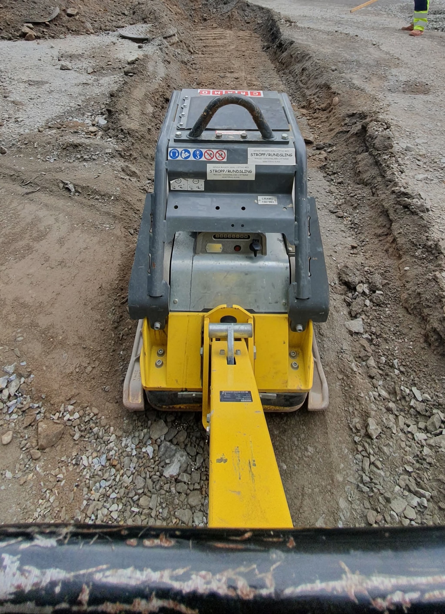 Żółto-szara zagęszczarka w wąskim wykopie, ubijająca podłoże. Widoczne ślady pracy i nierówności terenu. W tle fragment nogi pracownika w odblaskowym ubraniu.