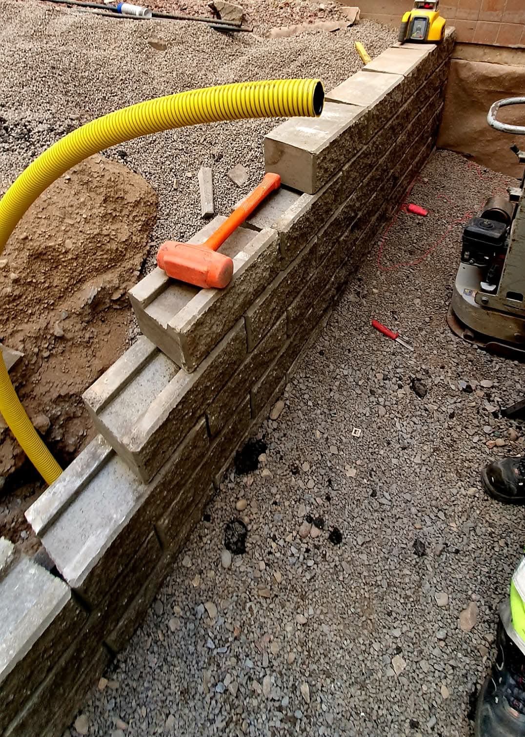 Budowa murku oporowego z betonowych bloczków, widoczna żółta rura drenażowa, pomarańczowy młotek i zagęszczarka. Etap prac ziemnych i przygotowania terenu.