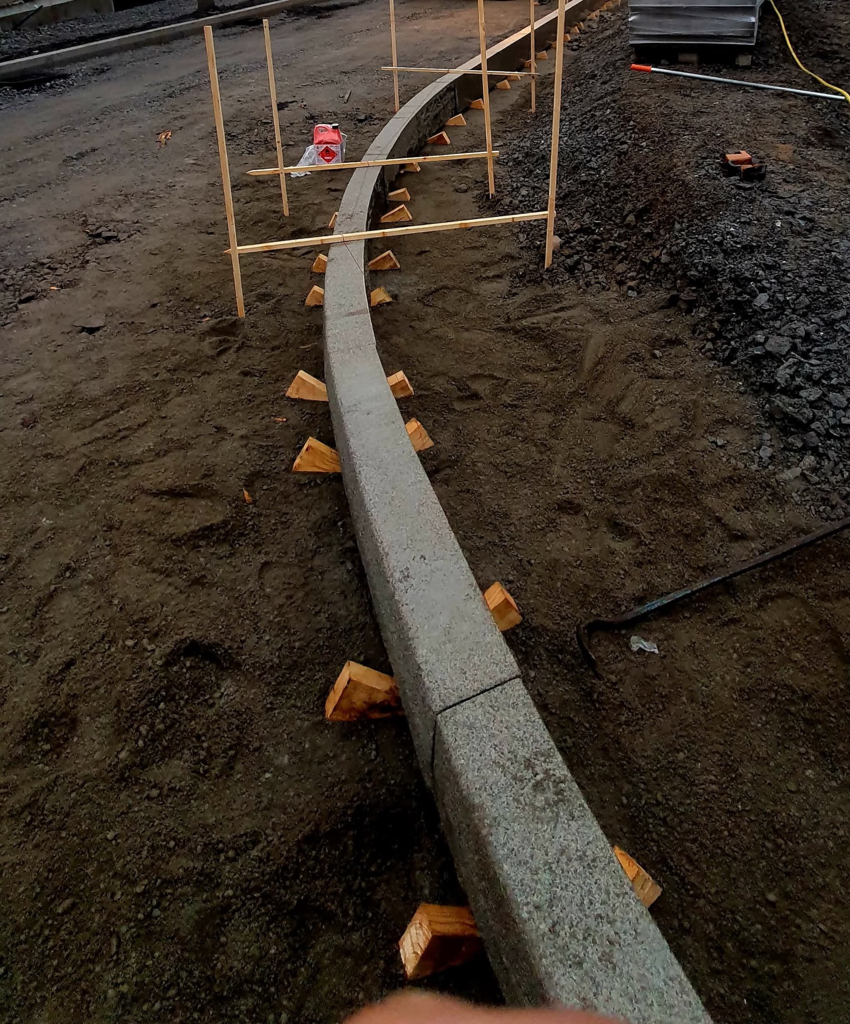 Zakrzywiony krawężnik z szarej kostki, podparty drewnianymi klinami i wyznaczony drewnianymi kołkami, na ziemi z gruzem. Etap prac brukarskich.