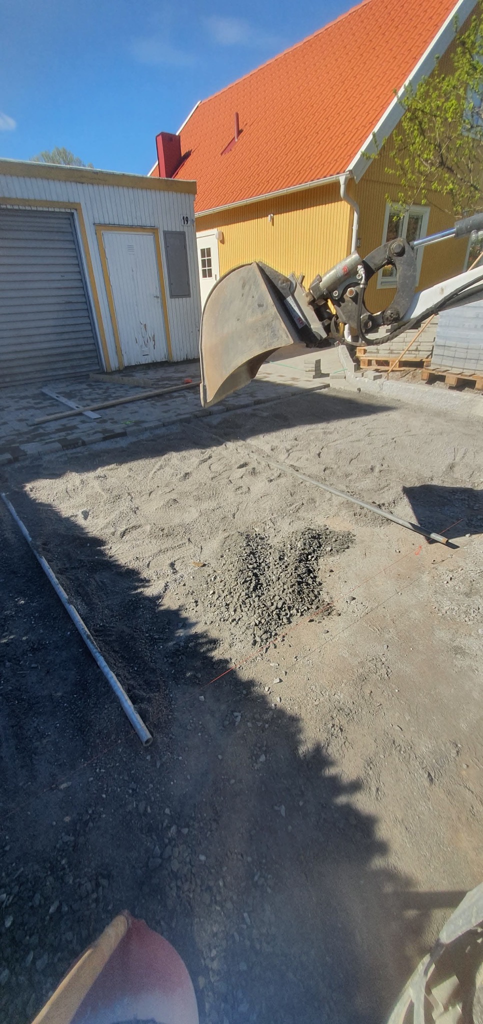 Praca koparką przy przygotowaniu podłoża pod kostkę brukową. Widoczny fragment ułożonej kostki, rura wyznaczająca poziom i żółty dom w tle. Słoneczny dzień.