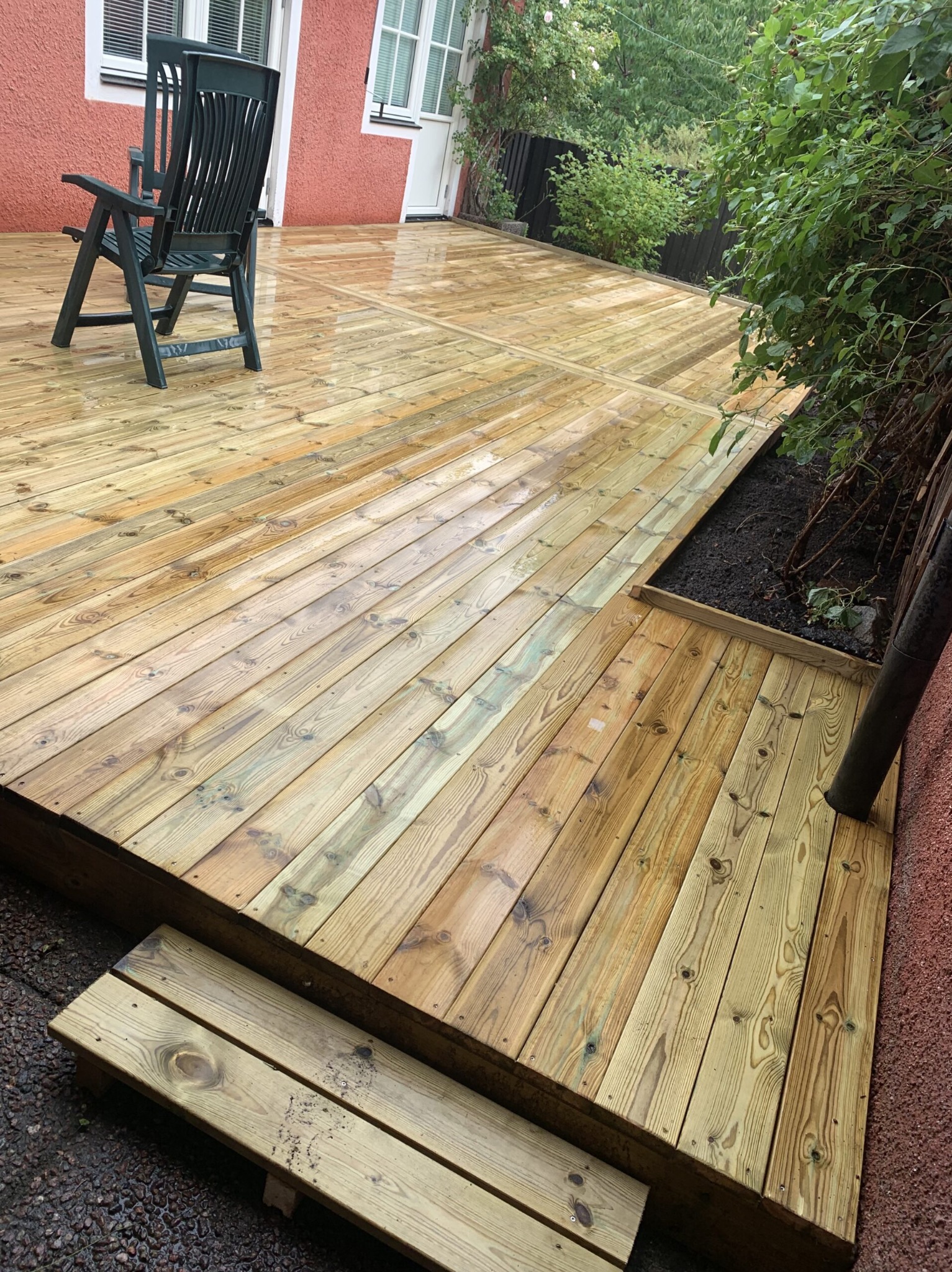 Świeżo wybudowany, drewniany taras z schodkami i krzesłem ogrodowym, po deszczu. Widoczne detale drewna i roślinność w tle.