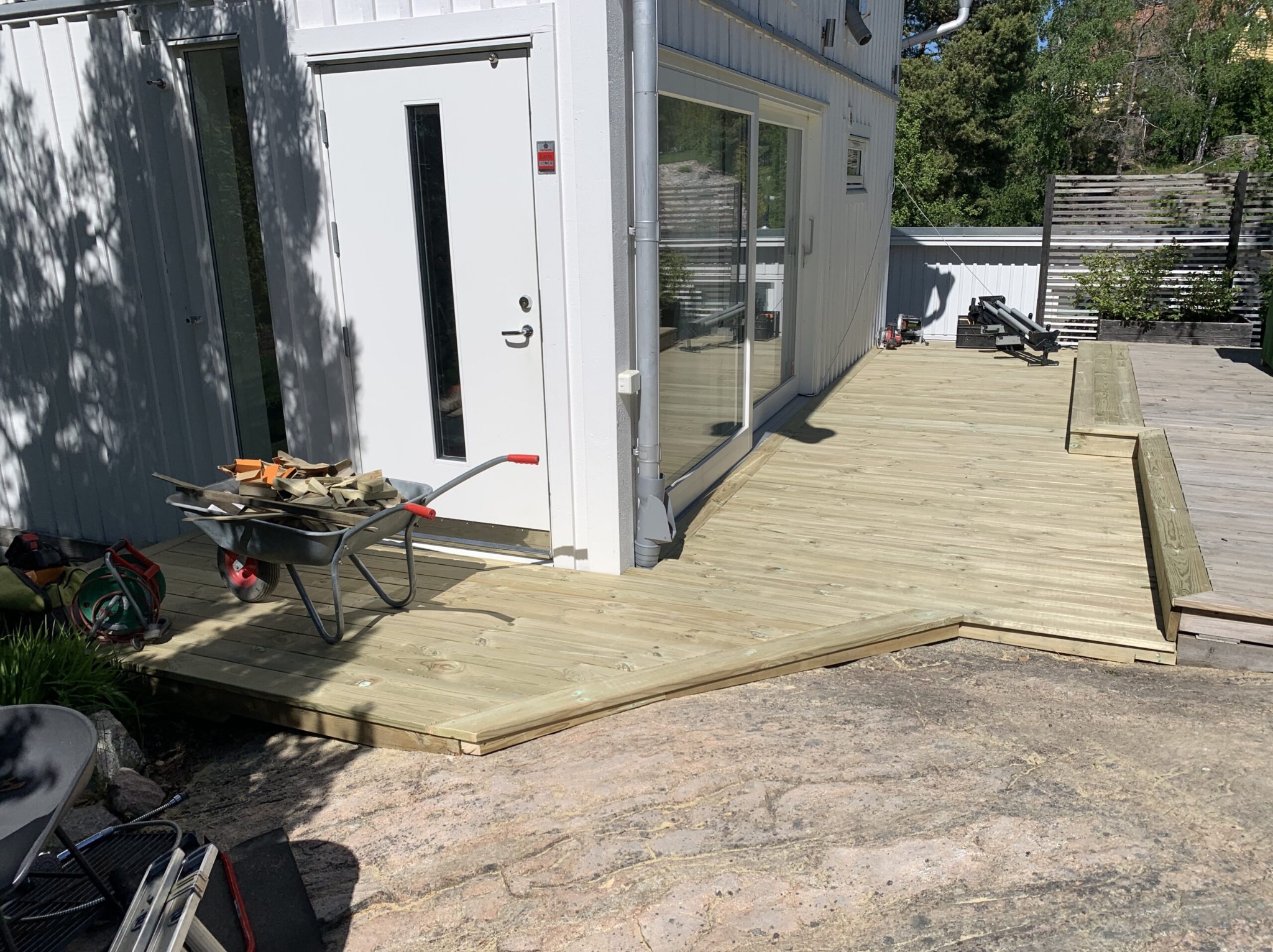 Nowy, drewniany taras przylegający do białego domu z widocznymi narzędziami i taczką z materiałami budowlanymi, położony na skalistym podłożu w słoneczny dzień.