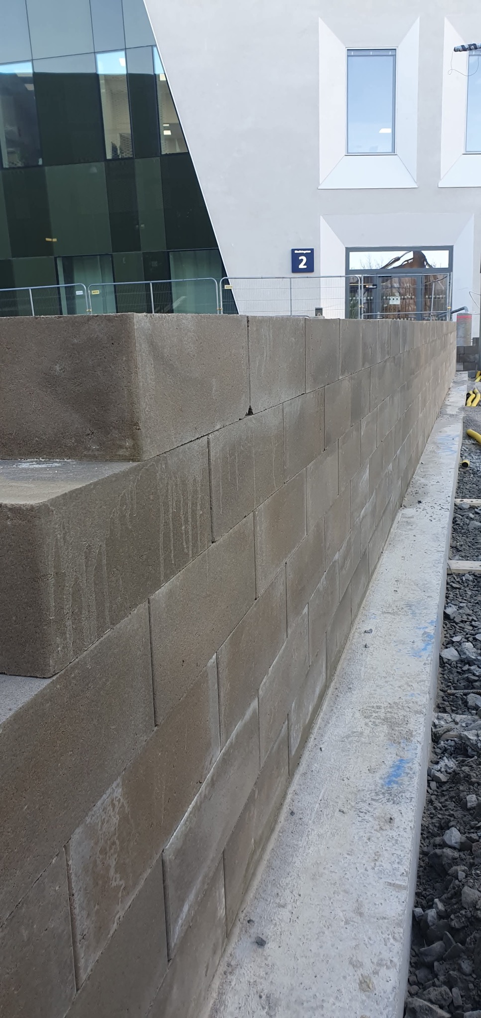 Świeżo wzniesiony murek z szarych bloczków betonowych, widoczny z boku, z fragmentem nowoczesnego budynku w tle. Widoczne ślady wilgoci na powierzchni bloczków.
