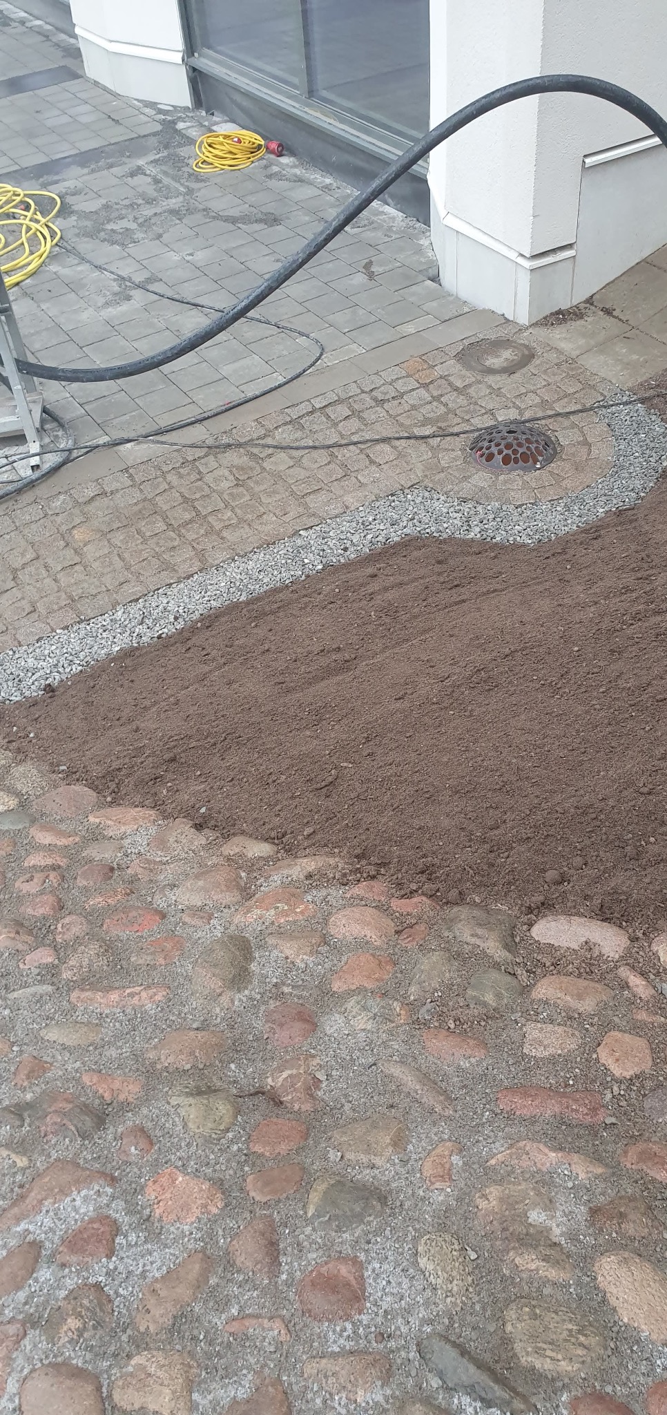 Fragment terenu z ułożoną kostką brukową, ziemią i kamieniami. Widoczny kabel i studzienka kanalizacyjna. Przygotowanie terenu pod ogród.