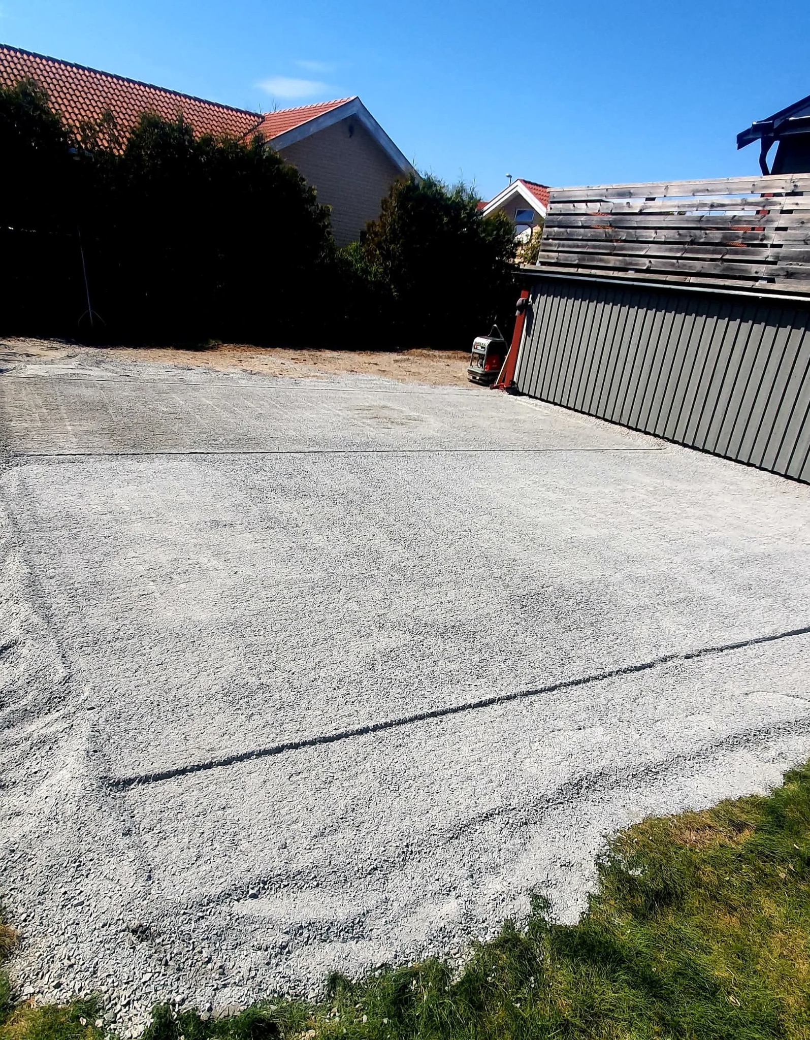 Przygotowany teren pod kostkę brukową z zagęszczarką w tle, widok z poziomu gruntu. Jasne, słoneczne oświetlenie.