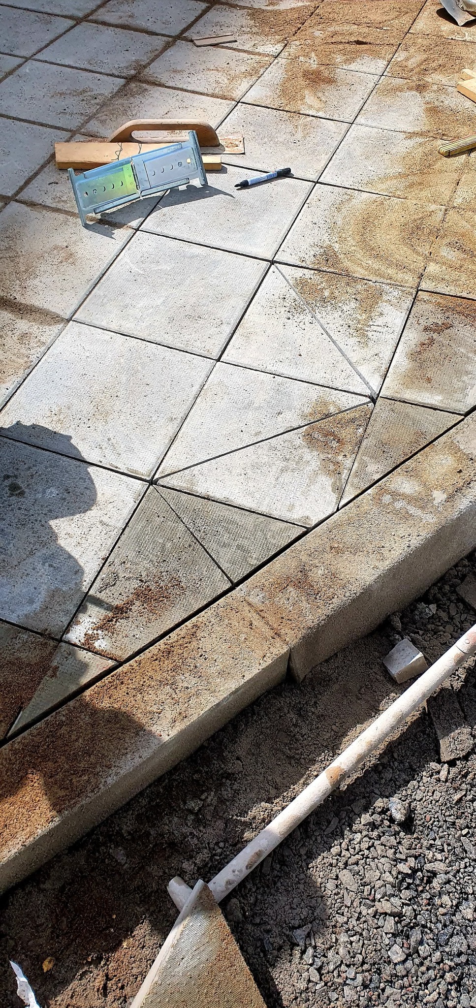 Układanie jasnoszarej kostki brukowej w geometryczny wzór. Widoczna poziomica, piasek i fragment łopaty. Krawężnik oddziela kostkę od gruntu.