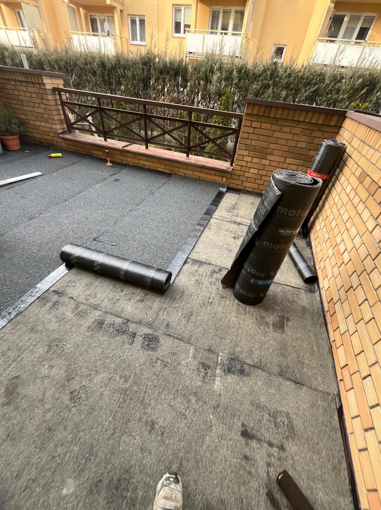 Częściowo położona izolacja dachu z papy bitumicznej, widoczne rolki materiału i fragment wykończonego dachu na tle ceglanego muru i zieleni.