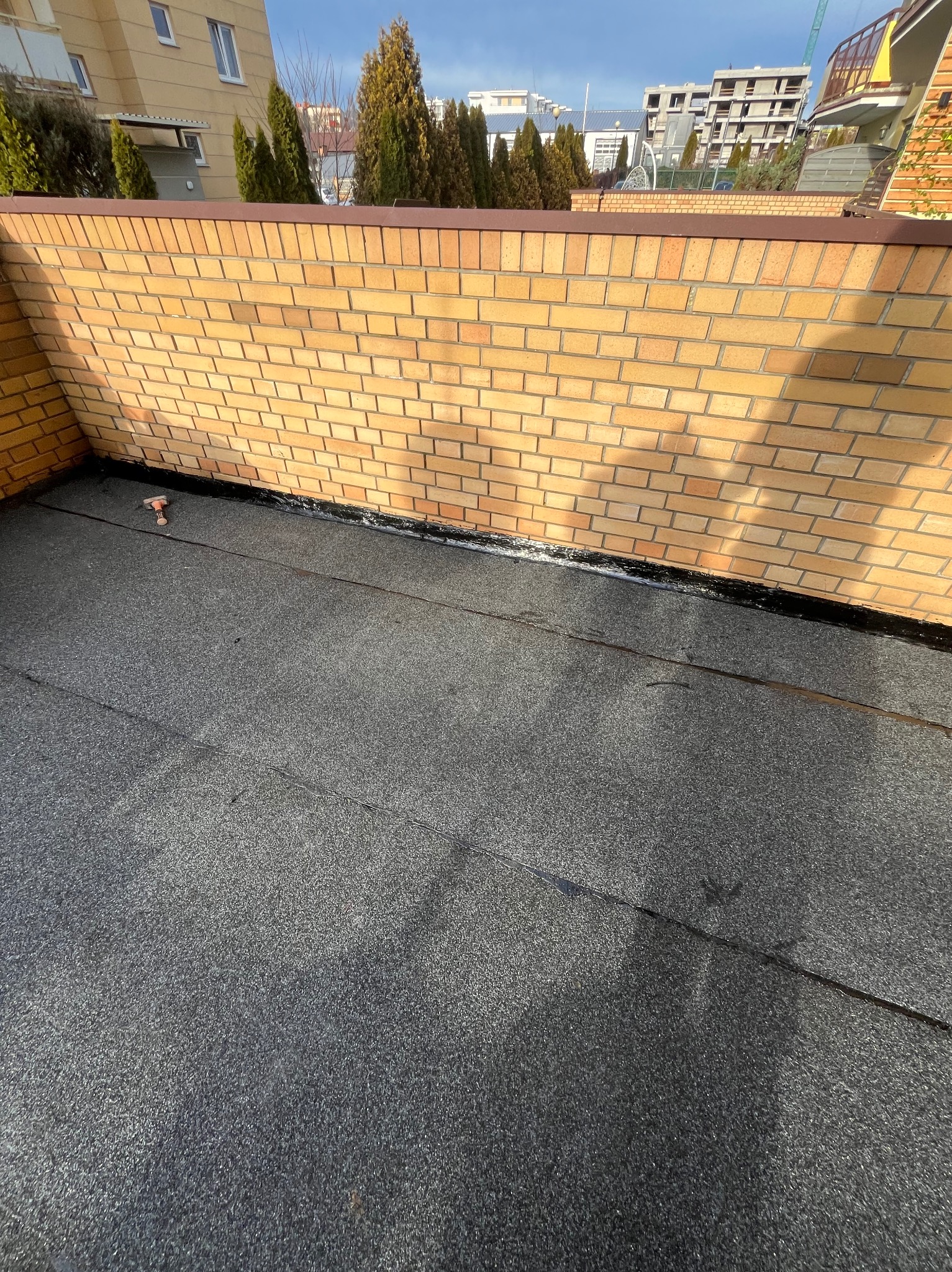 Szczelny dach pokryty papą na balkonie z ceglaną ścianą. Widoczne detale łączeń i narzędzie dekarskie. Tło stanowią budynki i drzewa.