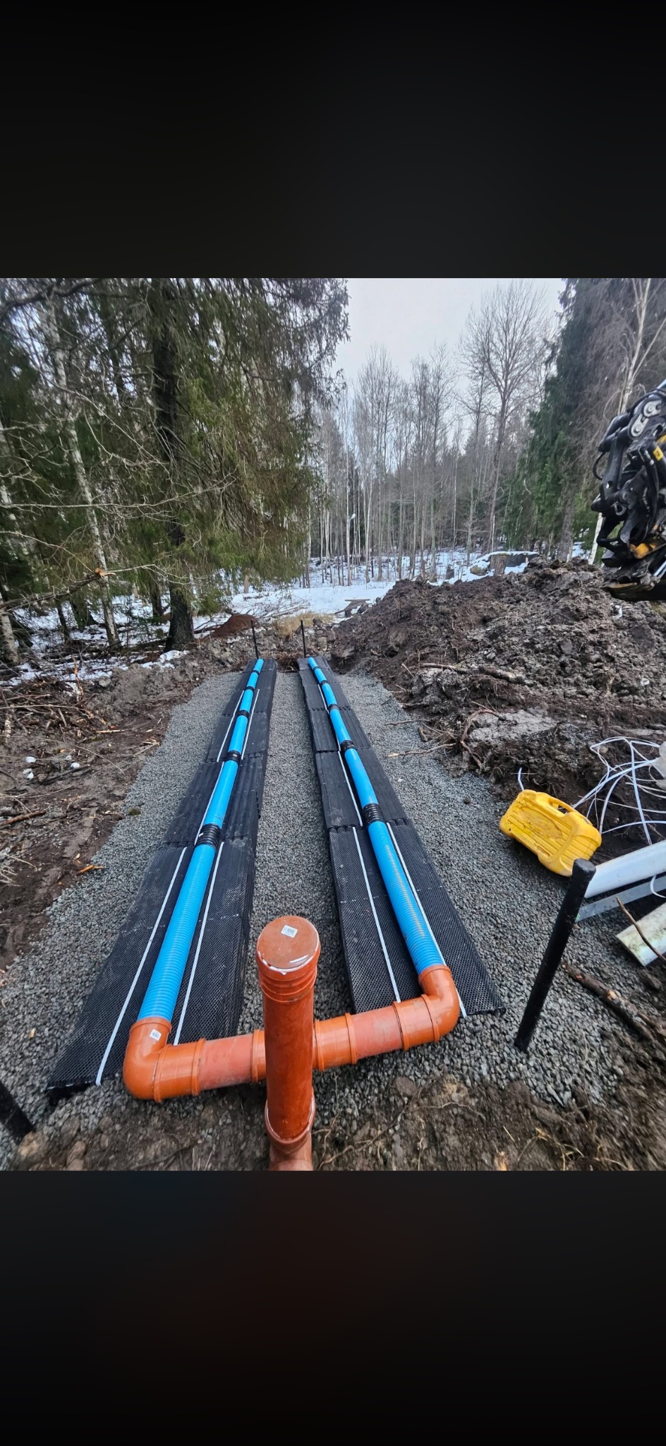 Drenaż liniowy z rurami w kolorze niebieskim i pomarańczowym, ułożony na czarnej macie drenażowej, na tle leśnego krajobrazu z resztkami śniegu.