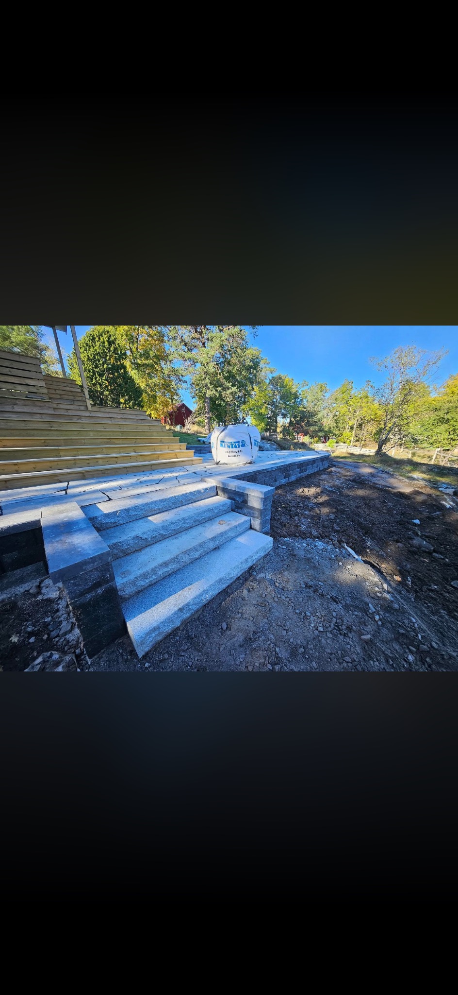 Granitowe schody zewnętrzne prowadzące do drewnianego tarasu, obok ziemia i worki z materiałem budowlanym, w tle zieleń i błękitne niebo.