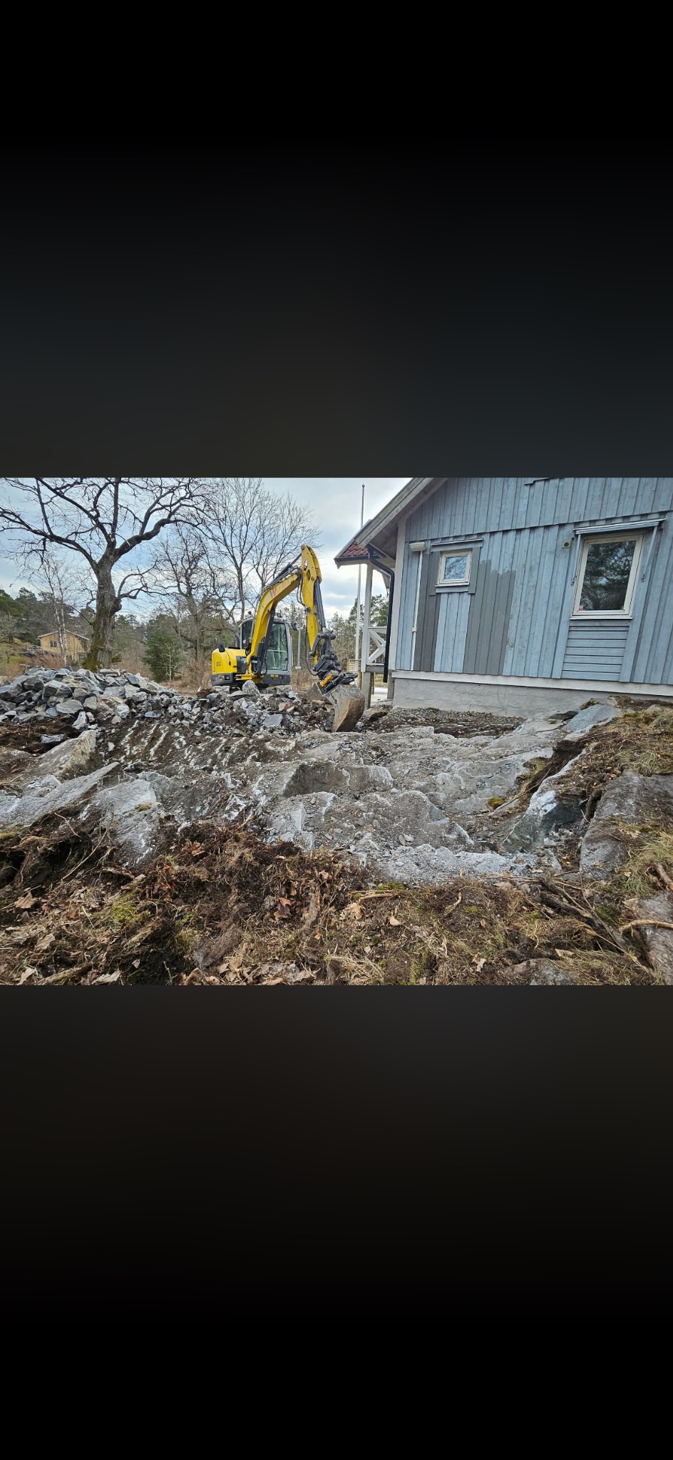 Żółta koparka na nierównym terenie skalnym obok szarego domu. Prace ziemne w toku, widoczne skały i ziemia. Drzewa w tle, pochmurne niebo.
