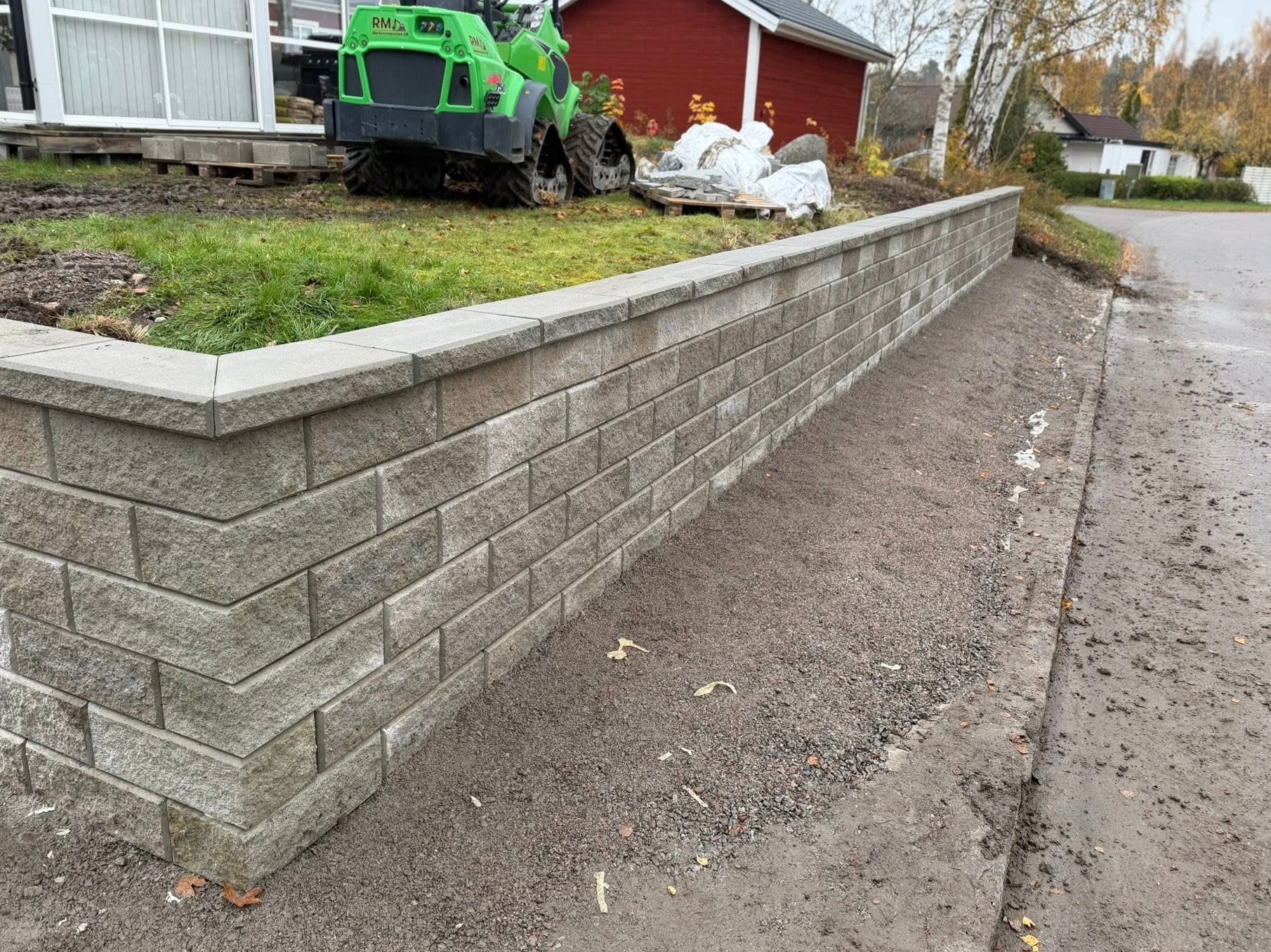 Nowo wybudowany murek oporowy z szarej kostki, zabezpieczający skarpę. W tle zielony miniładowarka i czerwony budynek. Praca wykonana w Szwecji.