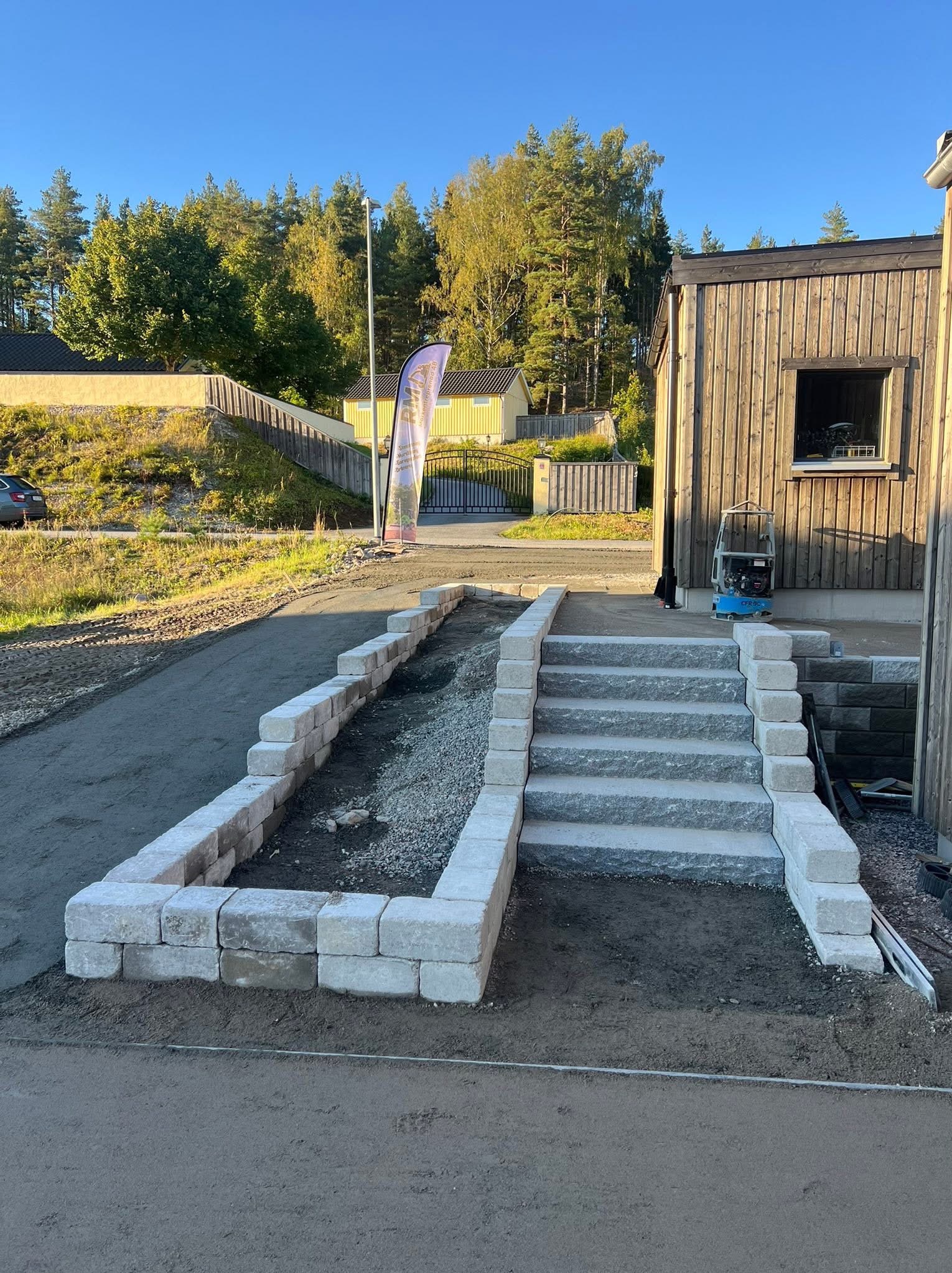 Ułożone schody z kostki brukowej prowadzące do domu, obok rabata z obrzeżem z kostki. W tle widoczny dom i drzewa w słoneczny dzień.