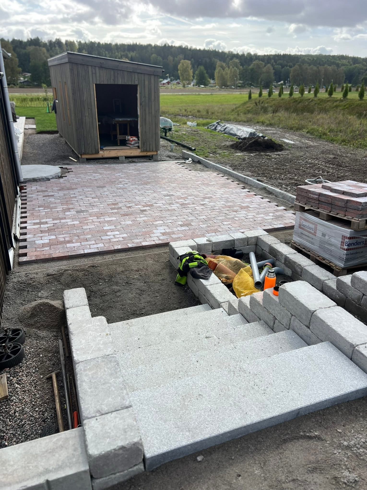 Częściowo ułożona, czerwono-szara kostka brukowa na posesji z drewnianym domkiem narzędziowym w tle. Widoczne schody z szarej kostki i paleta z kostką.