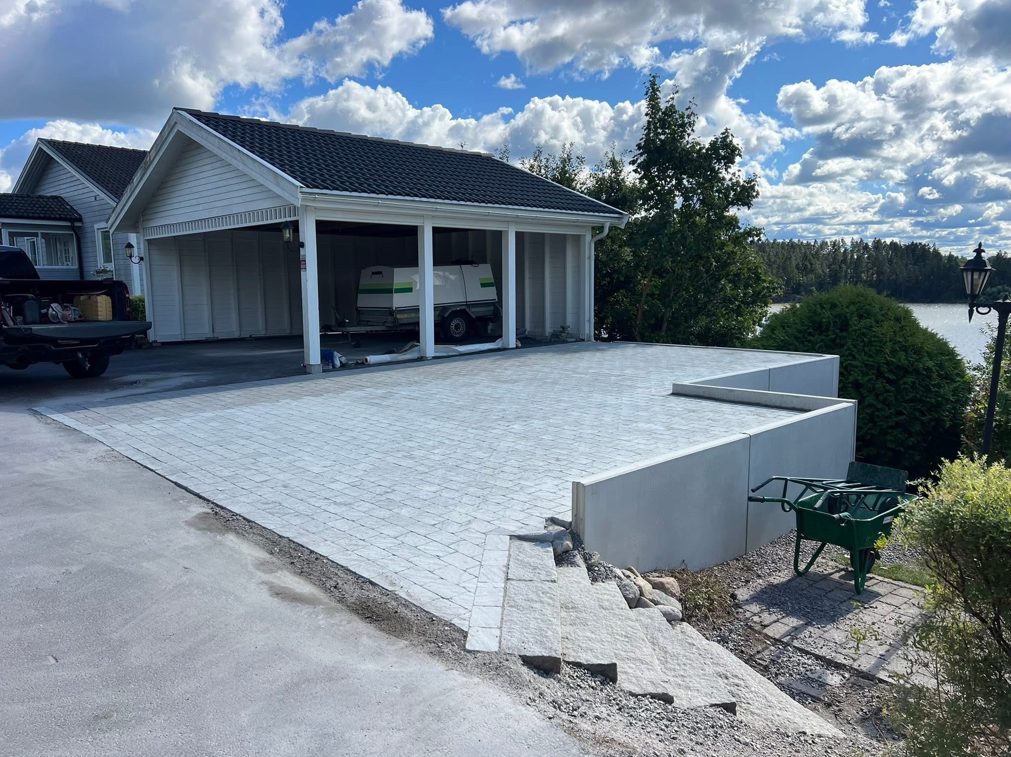 Jasnoszara kostka brukowa na podjeździe w Lublinie, prowadząca do garażu z przyczepą. Widok na jezioro i błękitne niebo z chmurami. W tle dom z ciemnym dachem.