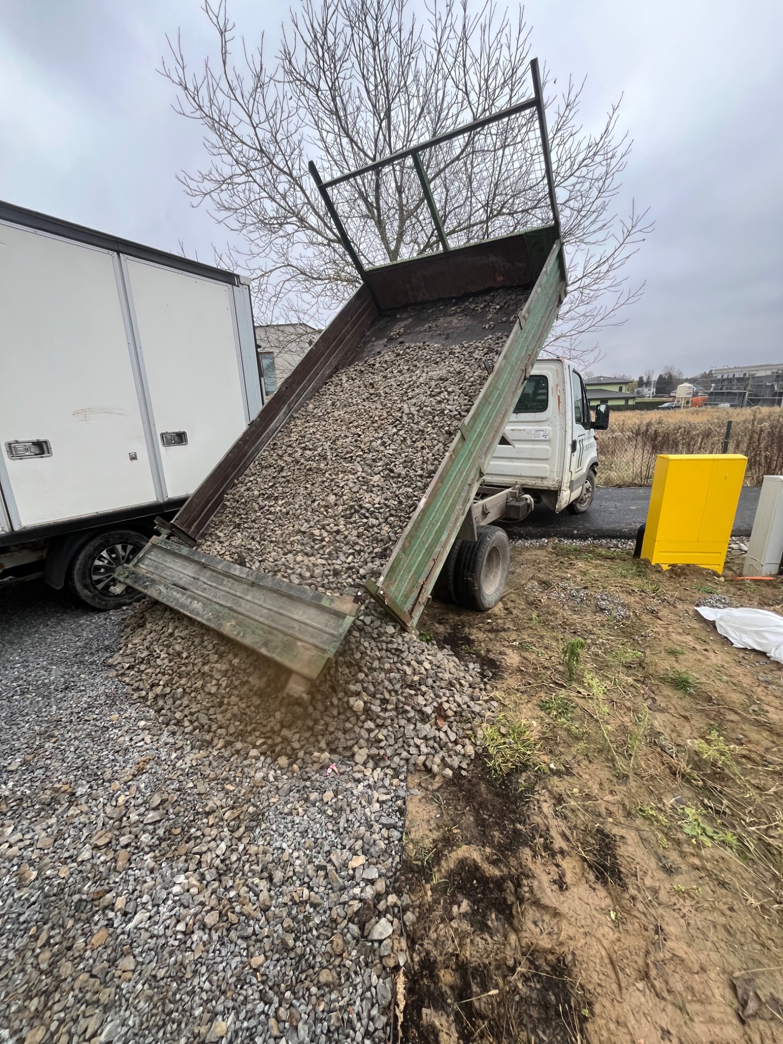 Wyładowany żwir z wywrotki na placu budowy, widoczny fragment białej naczepy i żółta skrzynka elektryczna. Szary, pochmurny dzień.