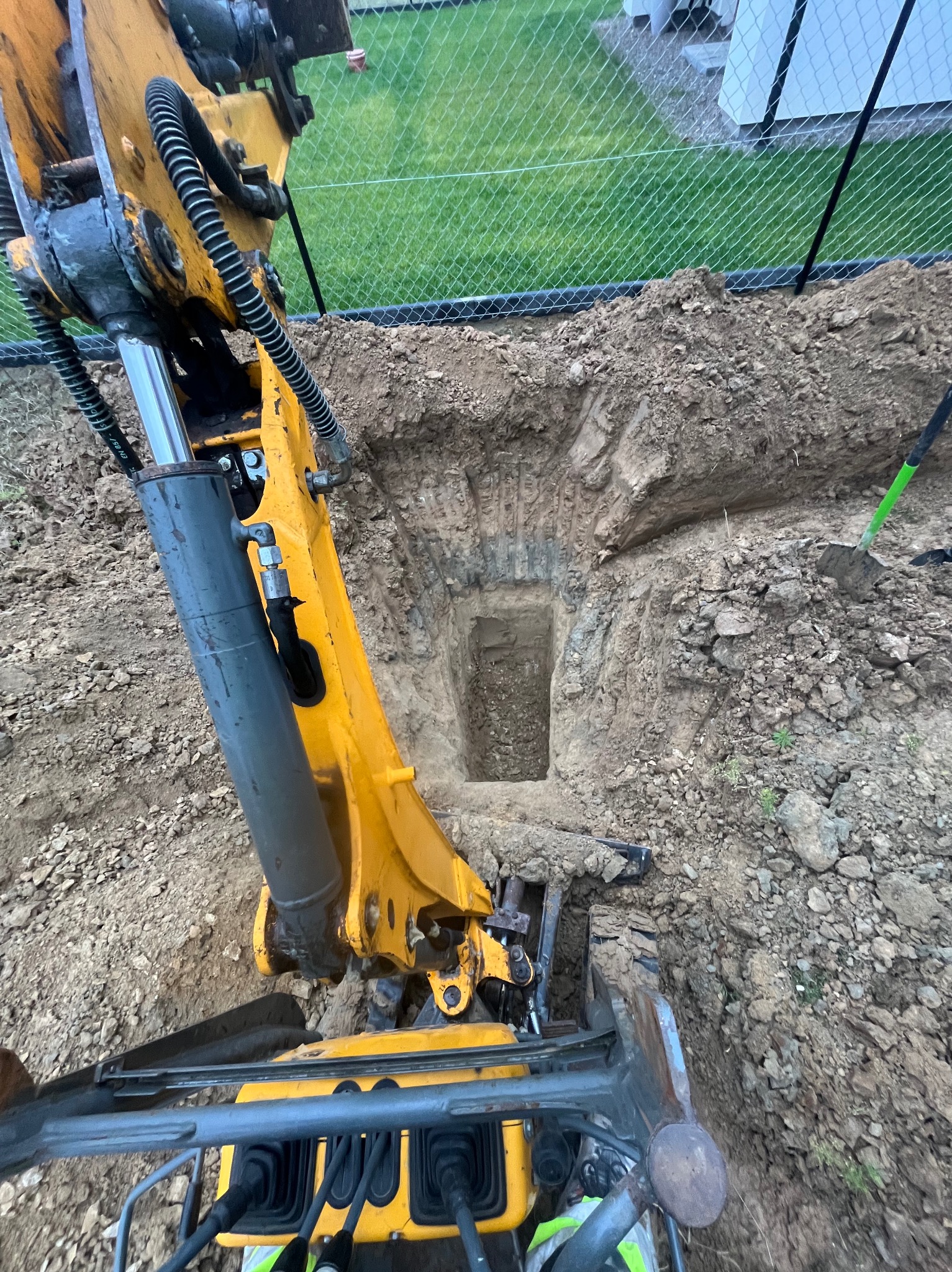 Żółta koparka w trakcie prac ziemnych, widoczny wykop w ziemi, w tle ogrodzenie z siatki. Ujęcie z góry na maszynę i wykop.
