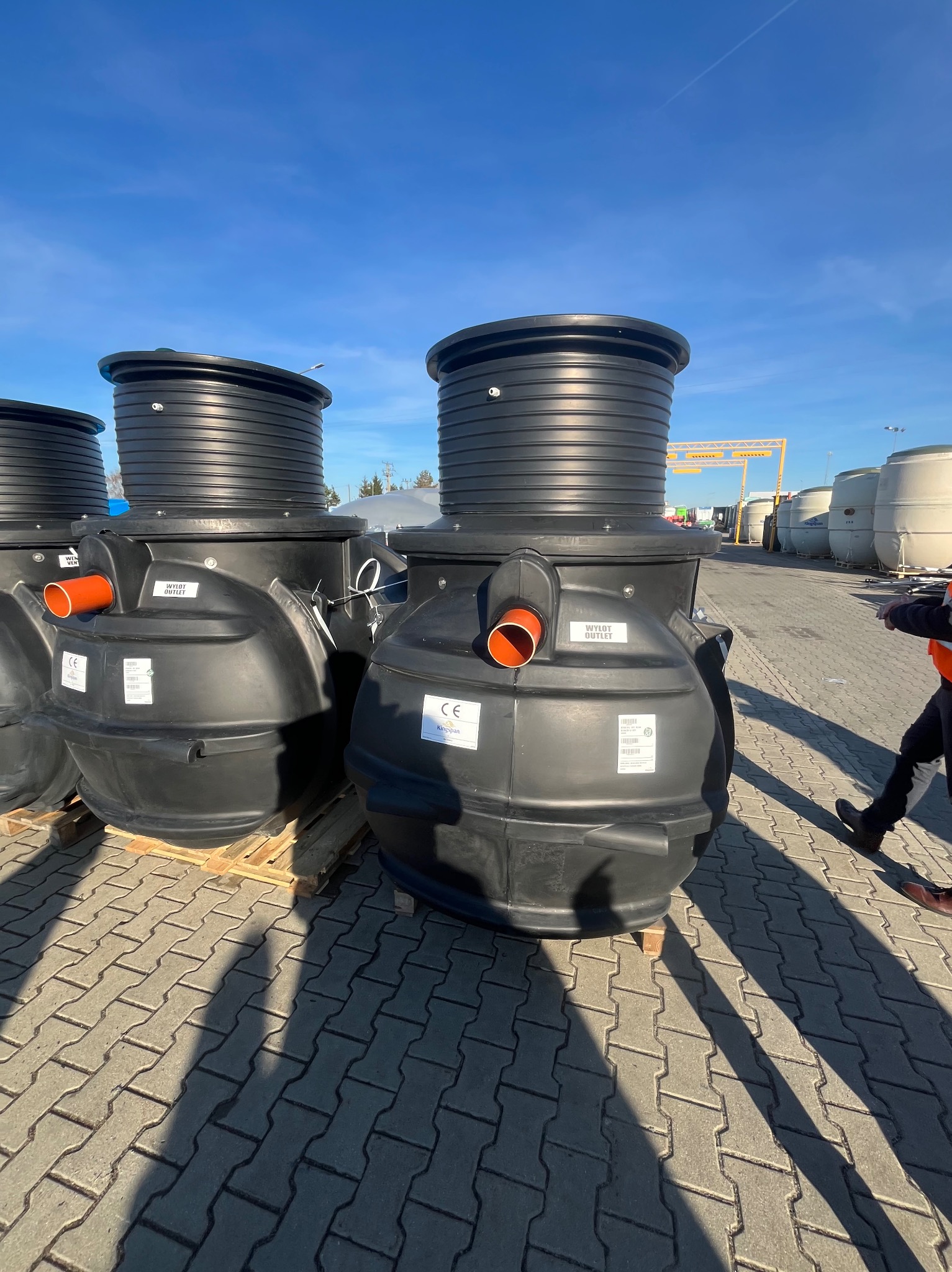 Czarne zbiorniki retencyjne z pomarańczowymi rurami wylotowymi, ustawione na kostce brukowej w słoneczny dzień. Widoczne etykiety informacyjne i fragment sylwetki osoby.