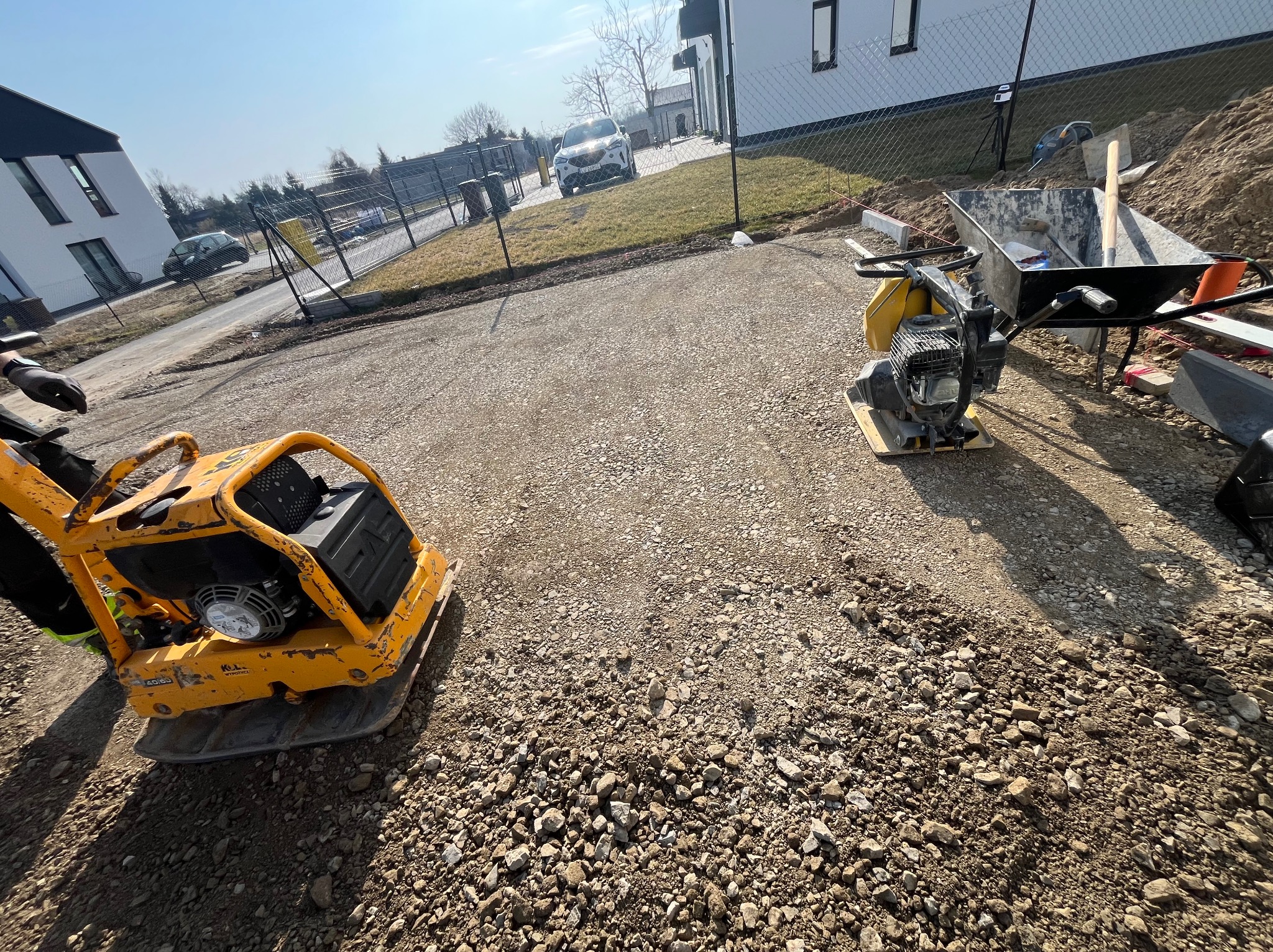 Żwirowa droga w trakcie utwardzania z użyciem zagęszczarki wibracyjnej. W tle widoczny dom jednorodzinny i ogrodzenie.