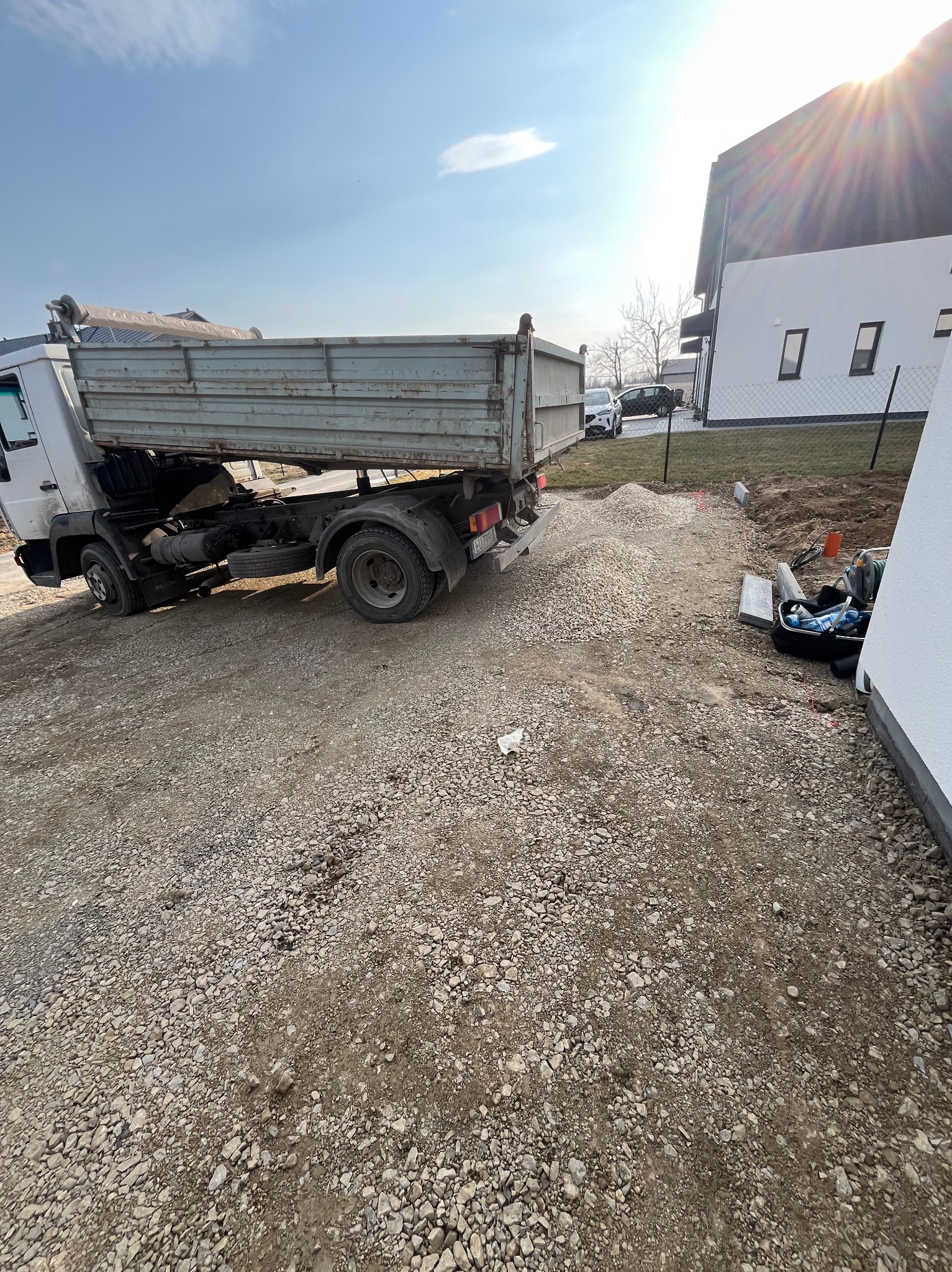 Wyładunek kruszywa z wywrotki na przygotowany teren pod budowę, widoczne narzędzia i materiały budowlane. Jasne niebo, nowoczesny dom w tle.