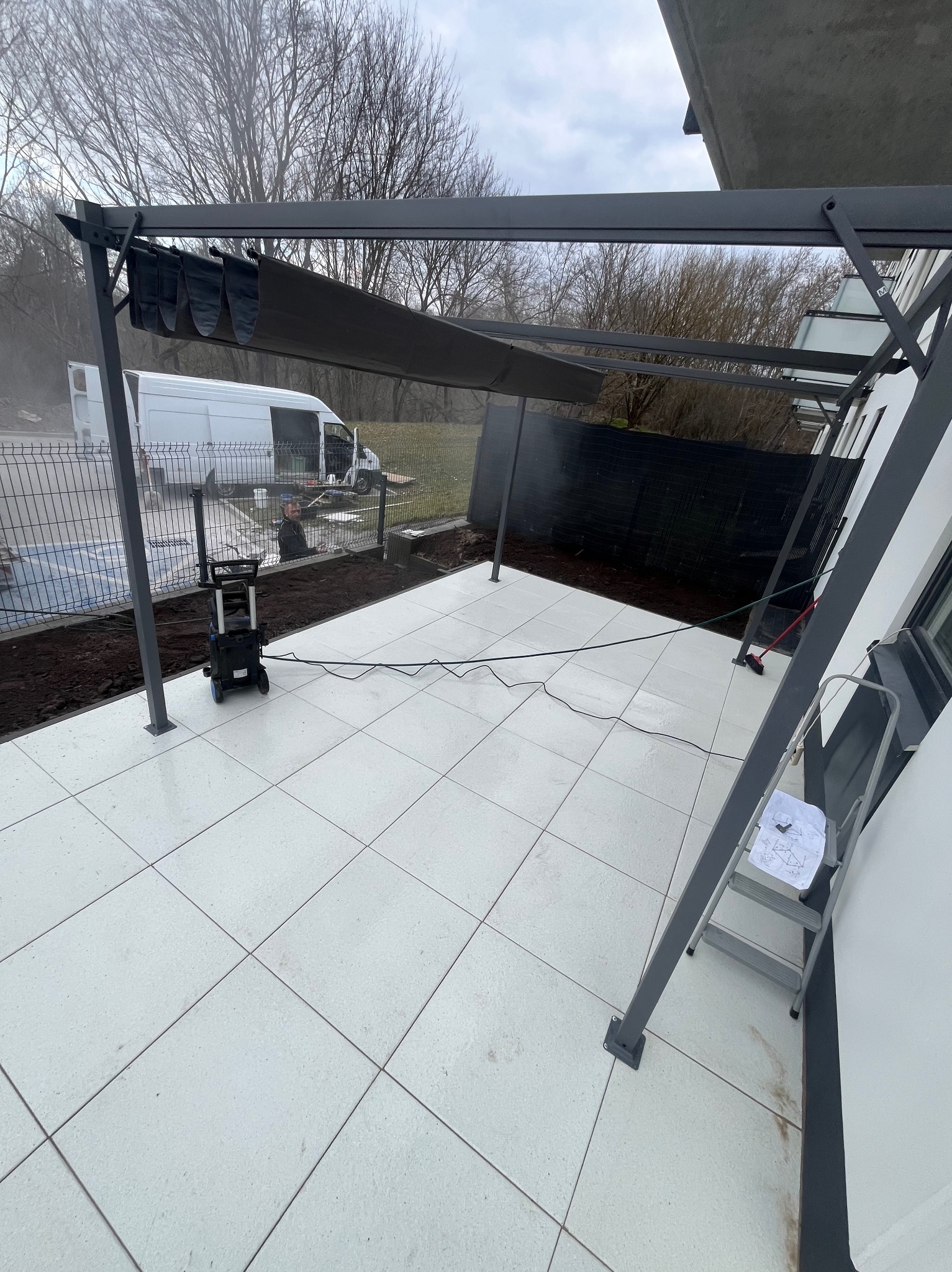 Nowoczesny taras z jasnych płyt, zadaszony pergolą w kolorze grafitowym. Widoczna myjka ciśnieniowa, drabina i częściowo zwinięte zadaszenie. W tle ogrodzenie i biały bus.