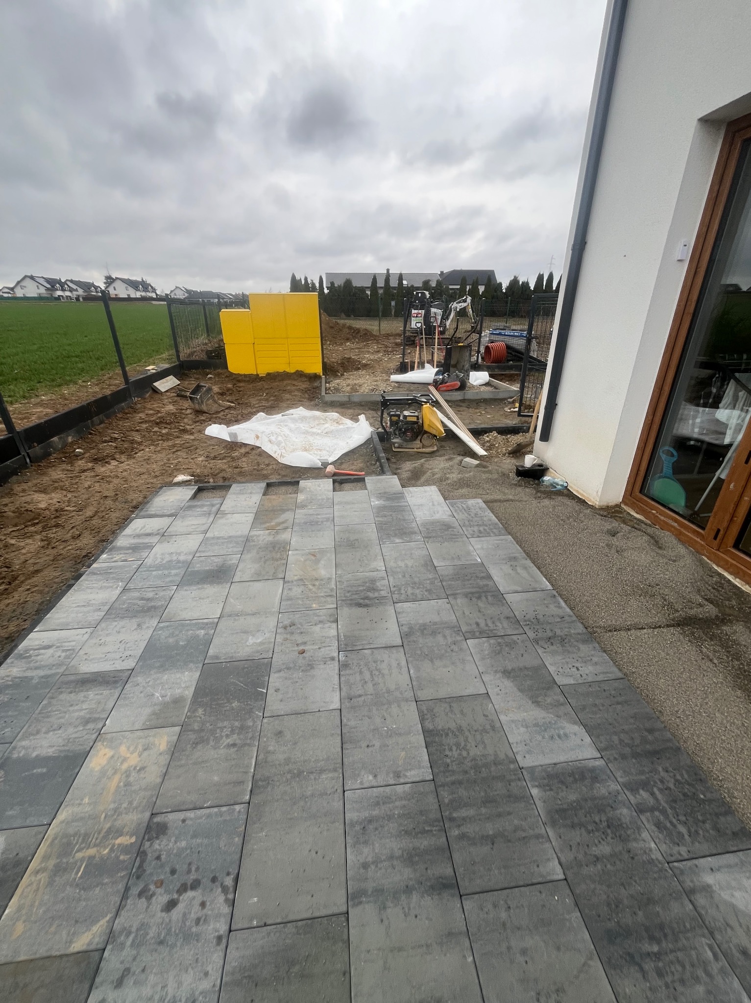Częściowo ułożona, szara kostka brukowa na posesji. W tle widoczne narzędzia, żółte płyty izolacyjne i dom z brązowymi oknami. Pochmurne niebo.