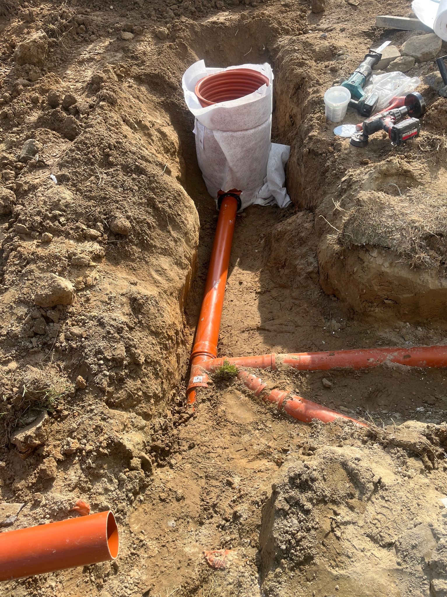 Pomarańczowe rury kanalizacyjne ułożone w wykopie ziemnym, widoczne narzędzia pracy. Etap budowy kanalizacji.