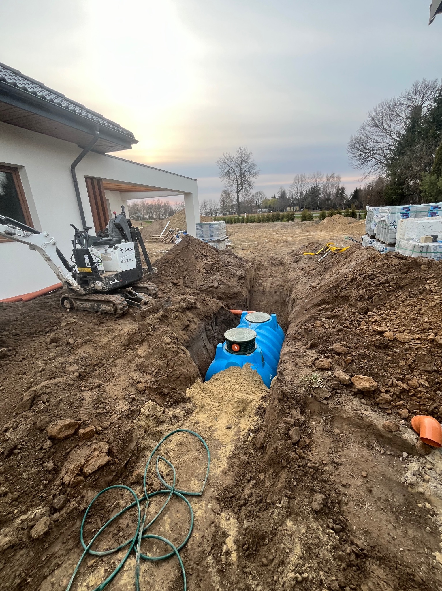 Niebieski zbiornik w wykopie, obok minikoparka Bobcat. W tle dom z jasną elewacją i sterta kostki brukowej. Prace ziemne w toku, widok z góry.