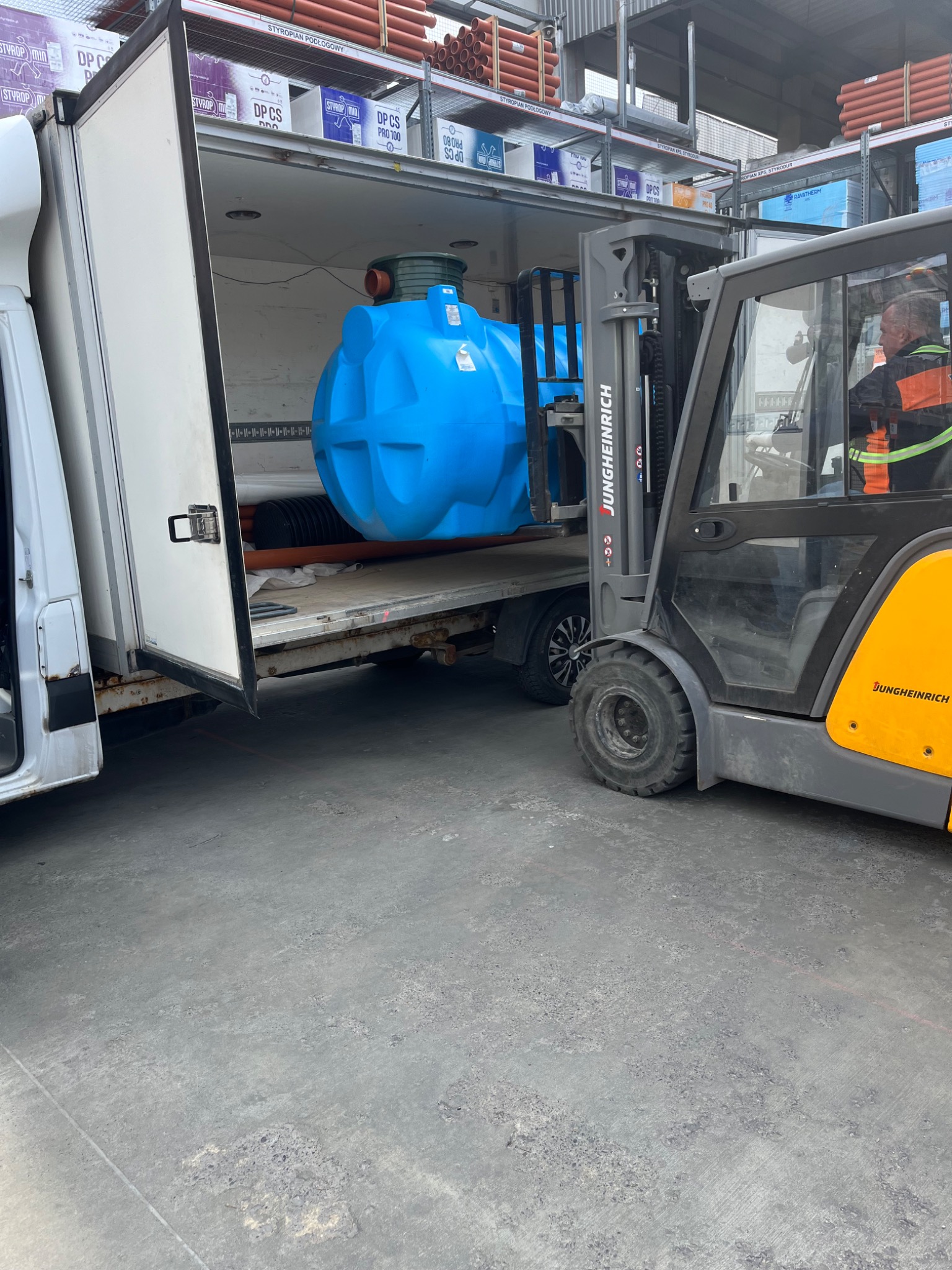 Wózek widłowy Jungheinrich ładuje niebieski zbiornik do białego busa. W tle widoczne materiały budowlane na regałach.
