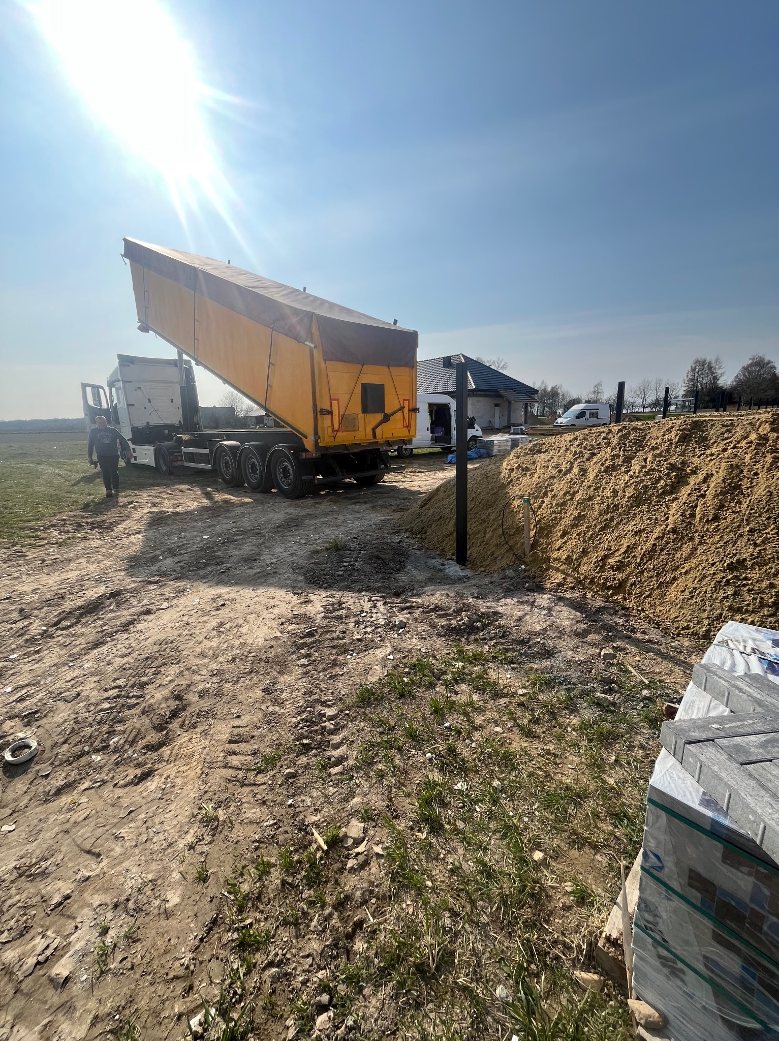 Żółta wywrotka z otwartą skrzynią obok sterty piasku i palety kostki brukowej na placu budowy w słoneczny dzień. W tle budynek i osoba.