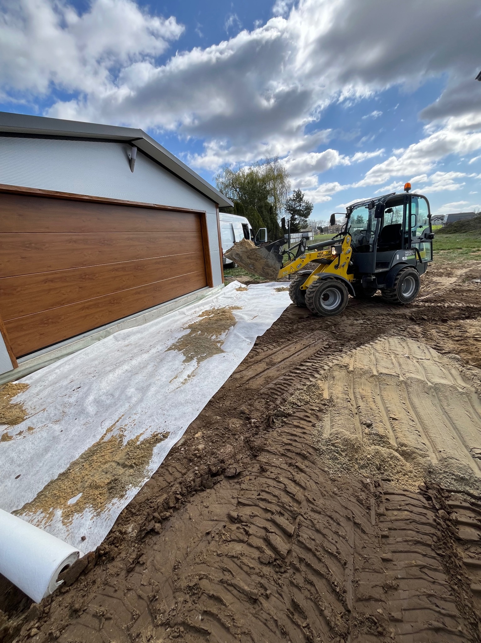 Żółta ładowarka Wacker Neuson na placu budowy, obok garażu z brązową bramą segmentową i rozłożoną geowłókniną. Widoczne ślady opon na ziemi.