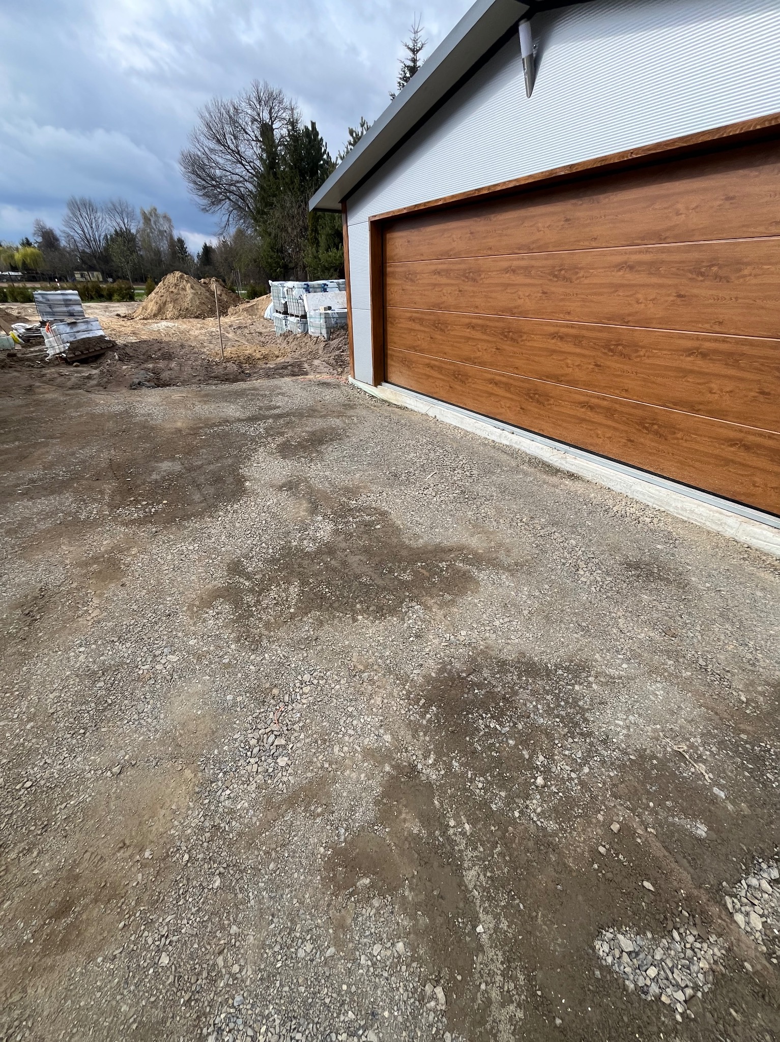 Przygotowany teren pod budowę z brązową bramą garażową i stertą kostki brukowej w tle, pochmurne niebo.