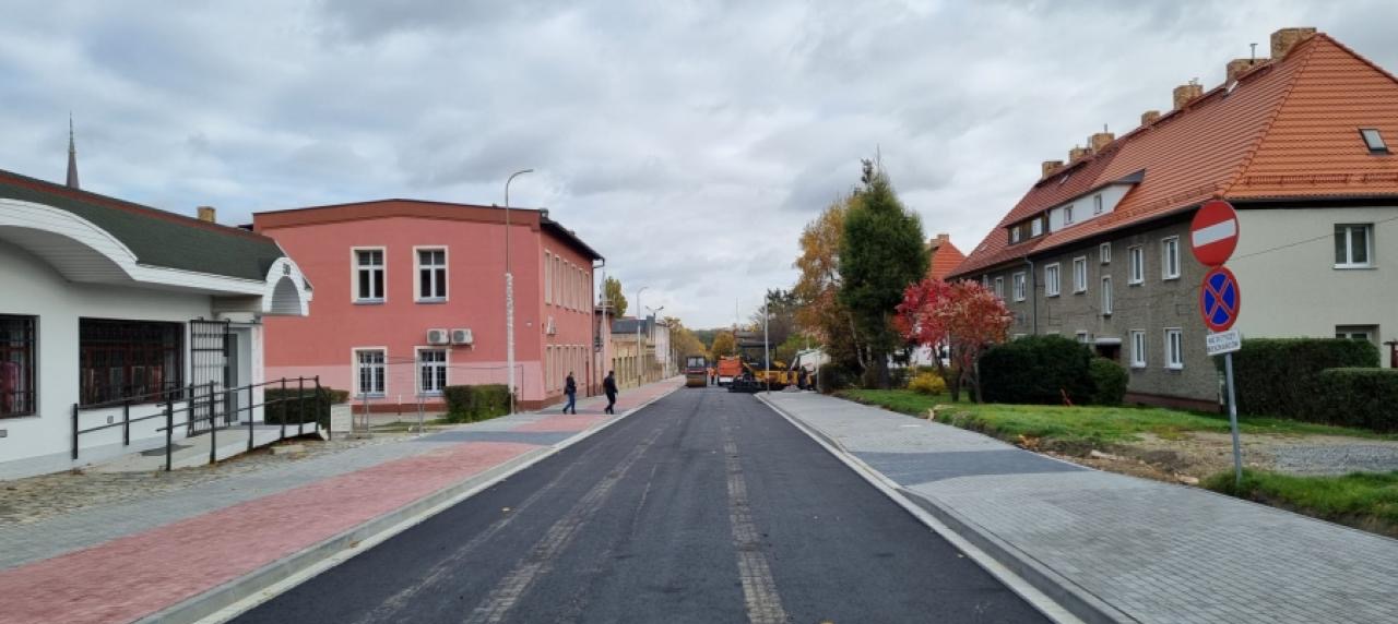Świeżo wyasfaltowana droga z kostką brukową po bokach, maszyny budowlane w oddali i budynki po obu stronach. Nowa nawierzchnia, chodniki z kostki, jesienne drzewa.